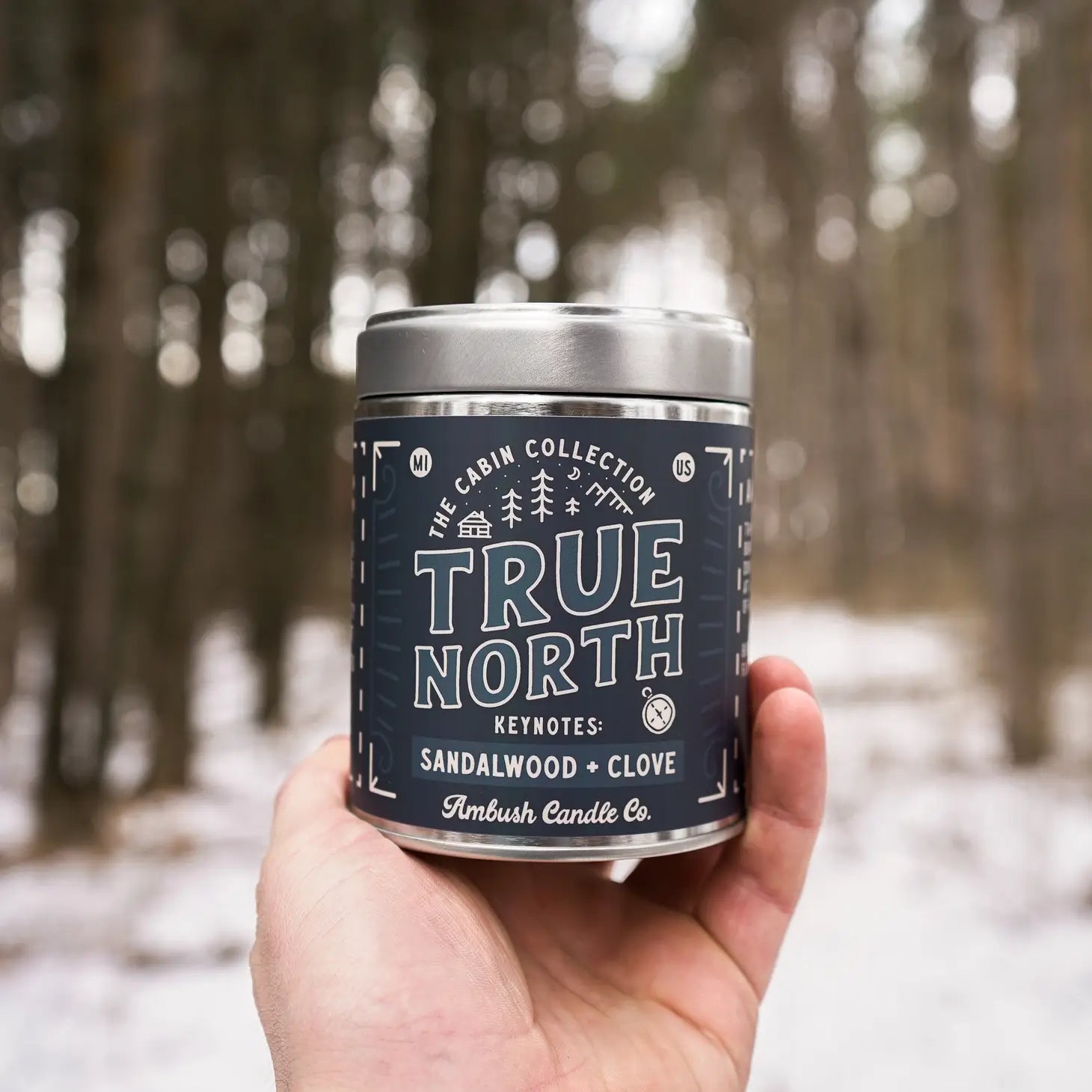 Hand holding an eco-friendly candle jar labeled "Ambush Candle Co 14OZ True North Sandalwood & Clove" in a snowy forest.