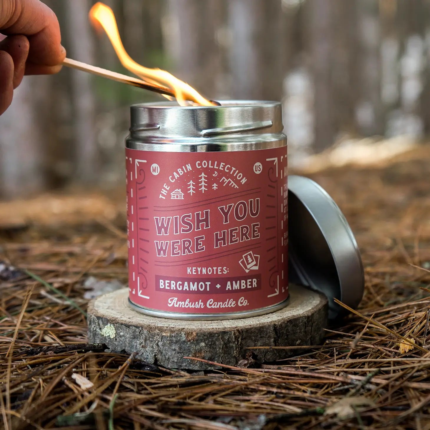 A hand with a lit match lights the "Ambush Candle Co 14OZ Wish You Were Here Bergamot & Amber Candle," made of clean-burning soy wax, placed on a tree stump.
