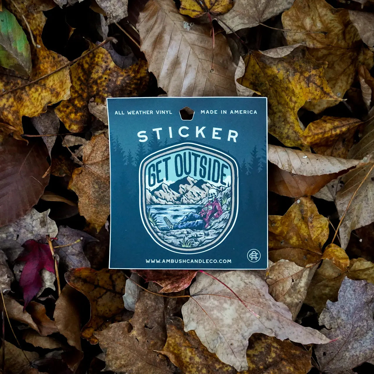 The Ambush Candle Co Get Outside Sticker, featuring the "GET OUTSIDE" text and a mountain scene, is placed among fallen autumn leaves. This weatherproof sticker, made in the USA, can be removed without damage.