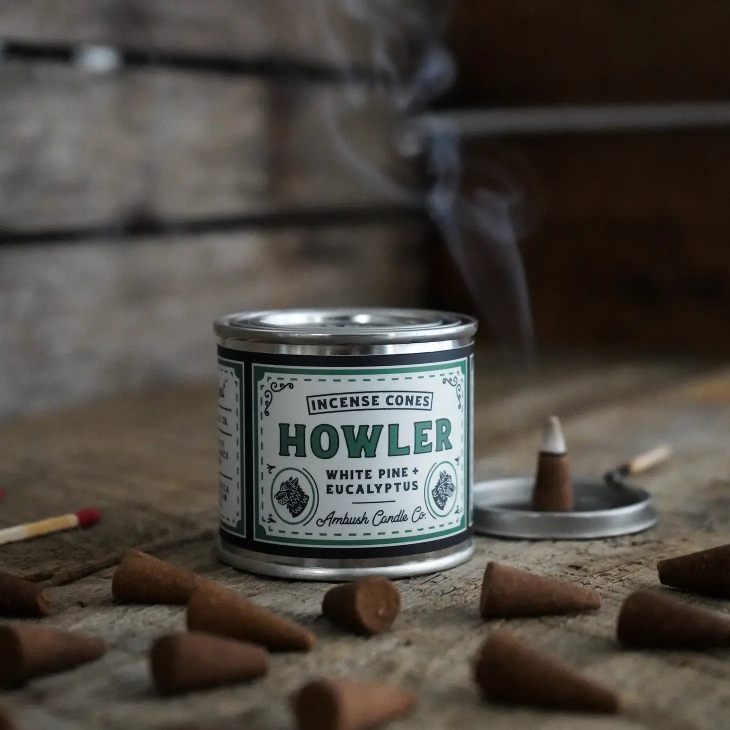 A tin of Ambush Candle Co Incense Cones in the White Pine & Eucalyptus - Howler scent, offering an invigorating eucalyptus fragrance and white pine aroma, surrounded by American-made incense cones with a smoking incense cone on a stand.
