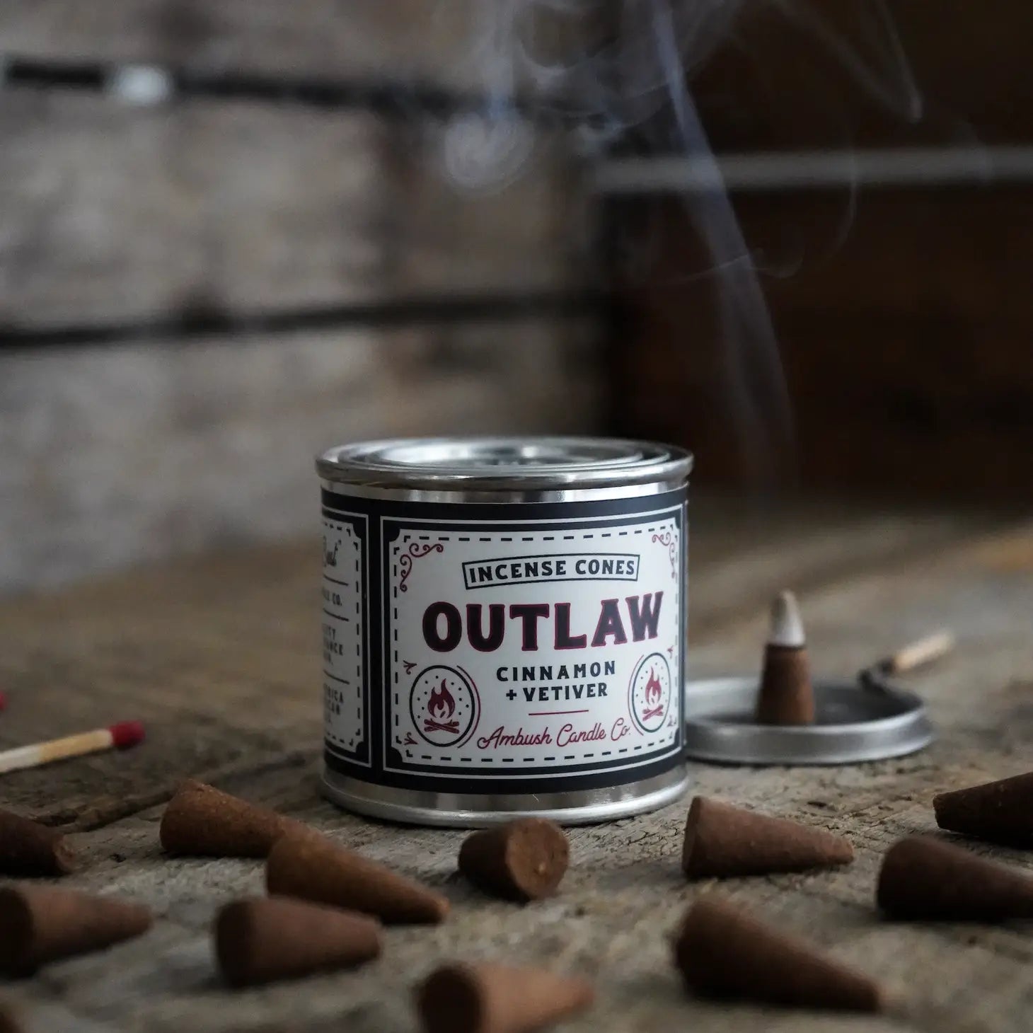 A tin labeled "Ambush Candle Co Incense Cones Cinnamon & Vetiver - Outlaw" with burning incense cones, matchsticks, and smoke rising in the background. These American-made incense cones offer a 20+ minute burn-time for each delightfully aromatic experience.