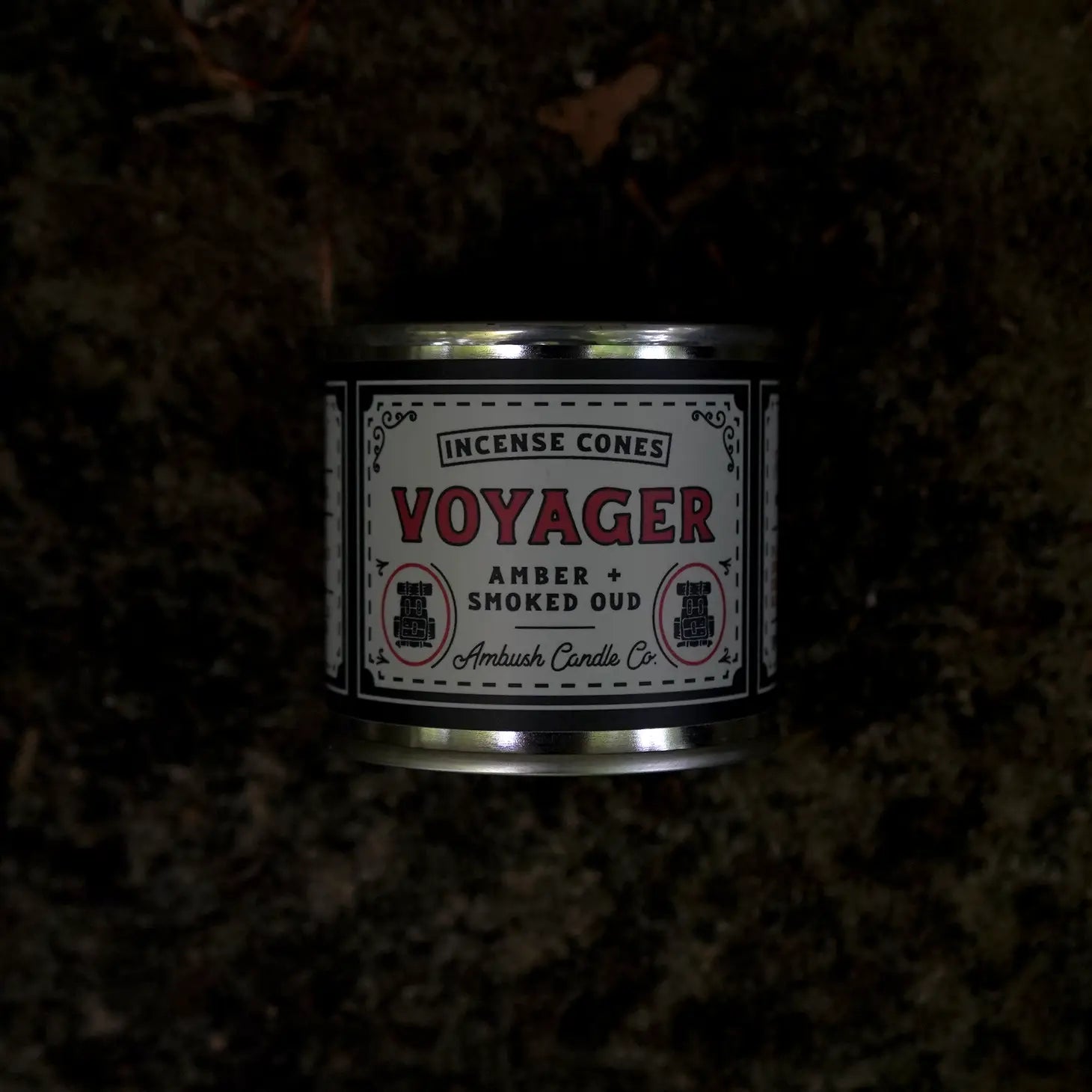 An exquisitely placed container of Ambush Candle Co Incense Cones, labeled "Amber + Smoked Oud - Voyager," rests on a dark, textured surface, emanating an air of mystery and sophistication.