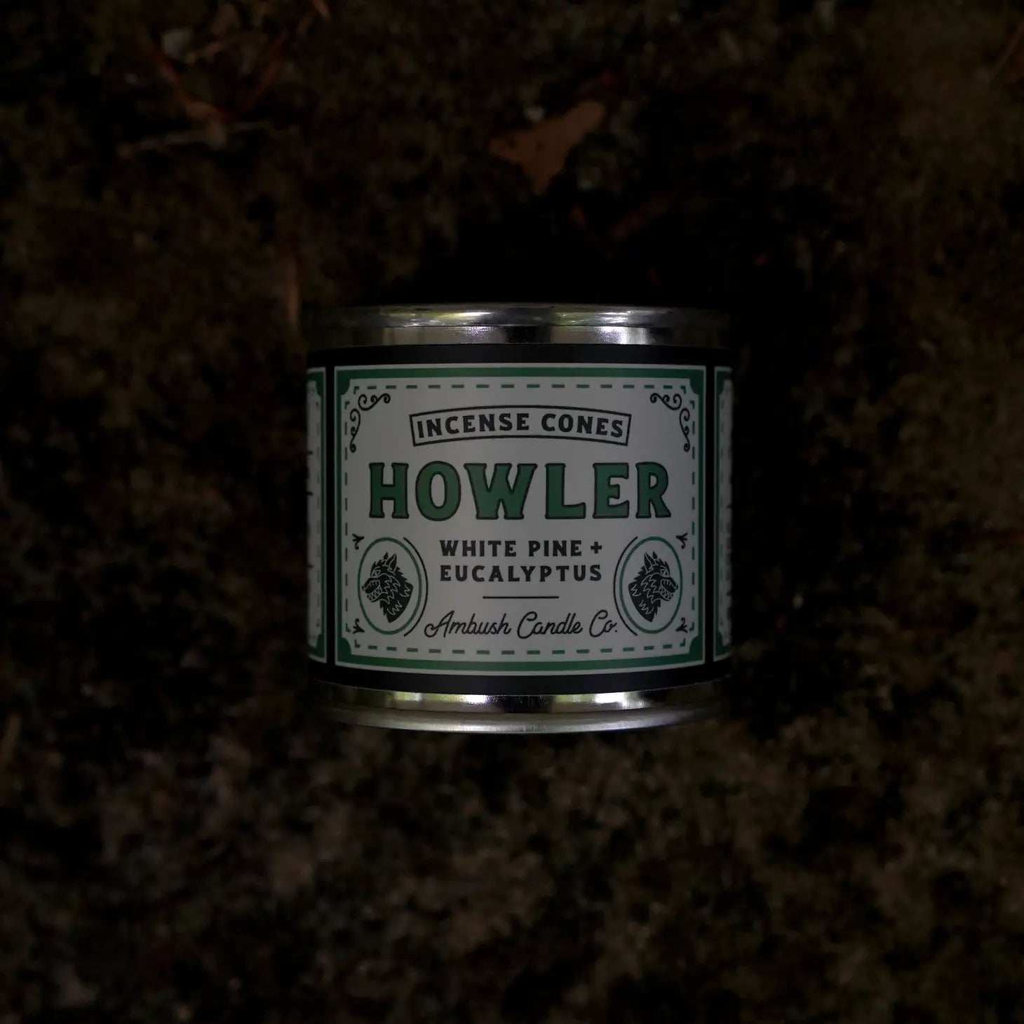 A tin of Ambush Candle Co Incense Cones in the White Pine & Eucalyptus - Howler scent, offering an invigorating eucalyptus fragrance and white pine aroma, surrounded by American-made incense cones with a smoking incense cone on a stand.