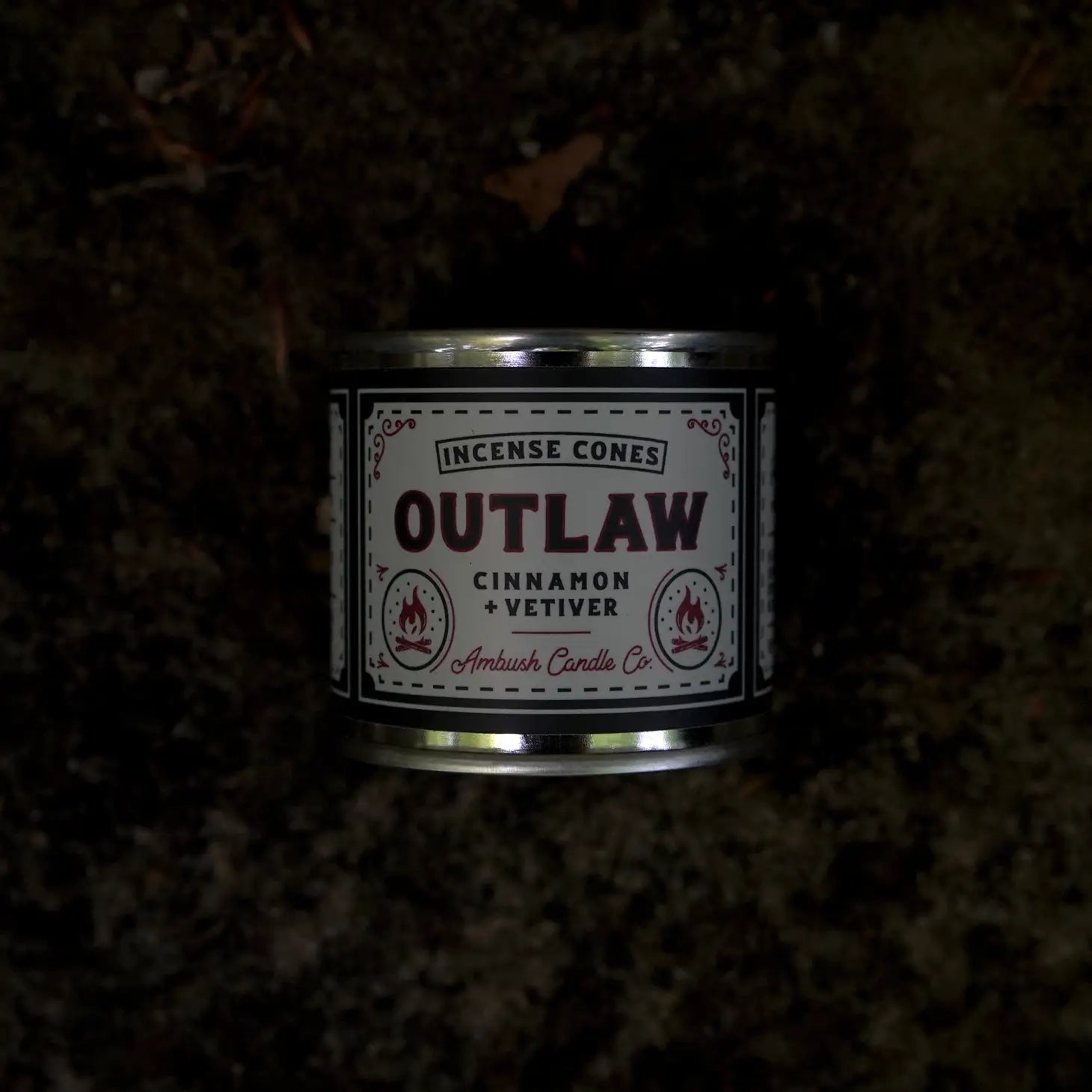 A tin labeled "Ambush Candle Co Incense Cones Cinnamon & Vetiver - Outlaw," American-made with a 20+ minute burn-time, is placed against a dark, textured background.