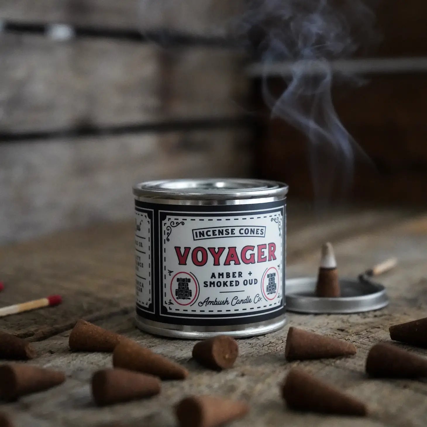 An exquisitely placed container of Ambush Candle Co Incense Cones, labeled "Amber + Smoked Oud - Voyager," rests on a dark, textured surface, emanating an air of mystery and sophistication.