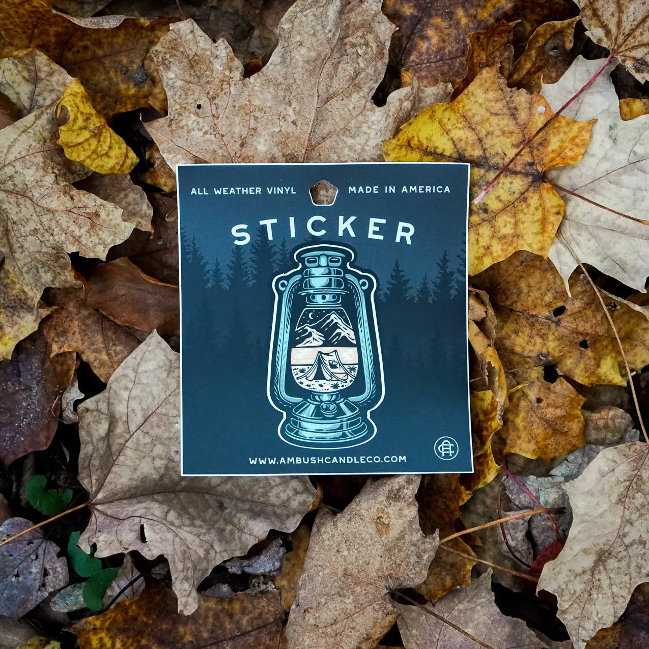 Introducing the Ambush Candle Co Lantern Sticker, showcasing a captivating mountain scene within a lantern design, elegantly placed on a bed of fallen autumn leaves. Crafted from weatherproof vinyl, this removable sticker is perfect for adorning any surface. The text reads "Sticker" and "www.ambushcandleco.com".
