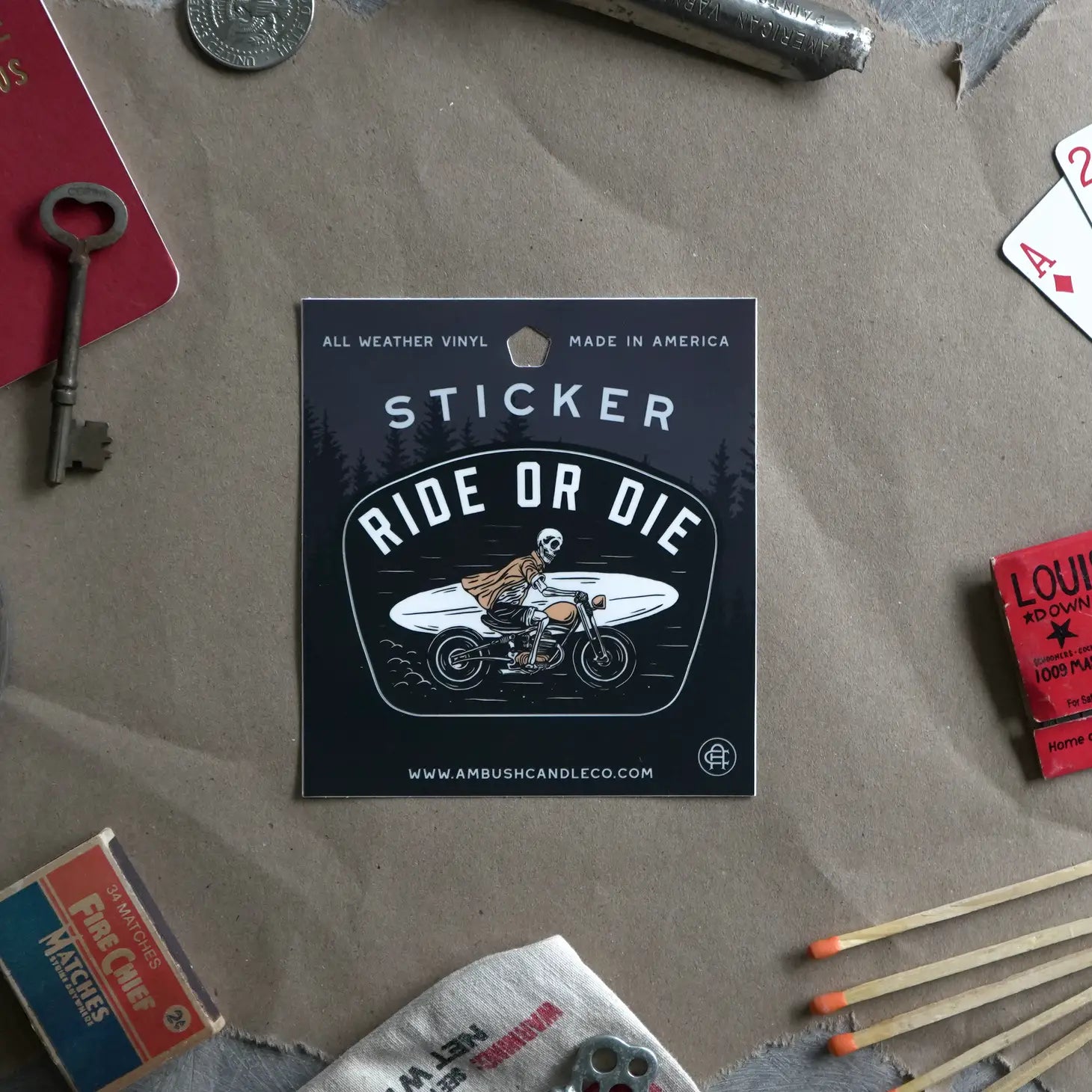 On a table, a weatherproof vinyl Ambush Candle Co "Ride or Die" sticker displays an image of a person on a bike, surrounded by various vintage items. This proudly made in the USA sticker adds character to the scene.