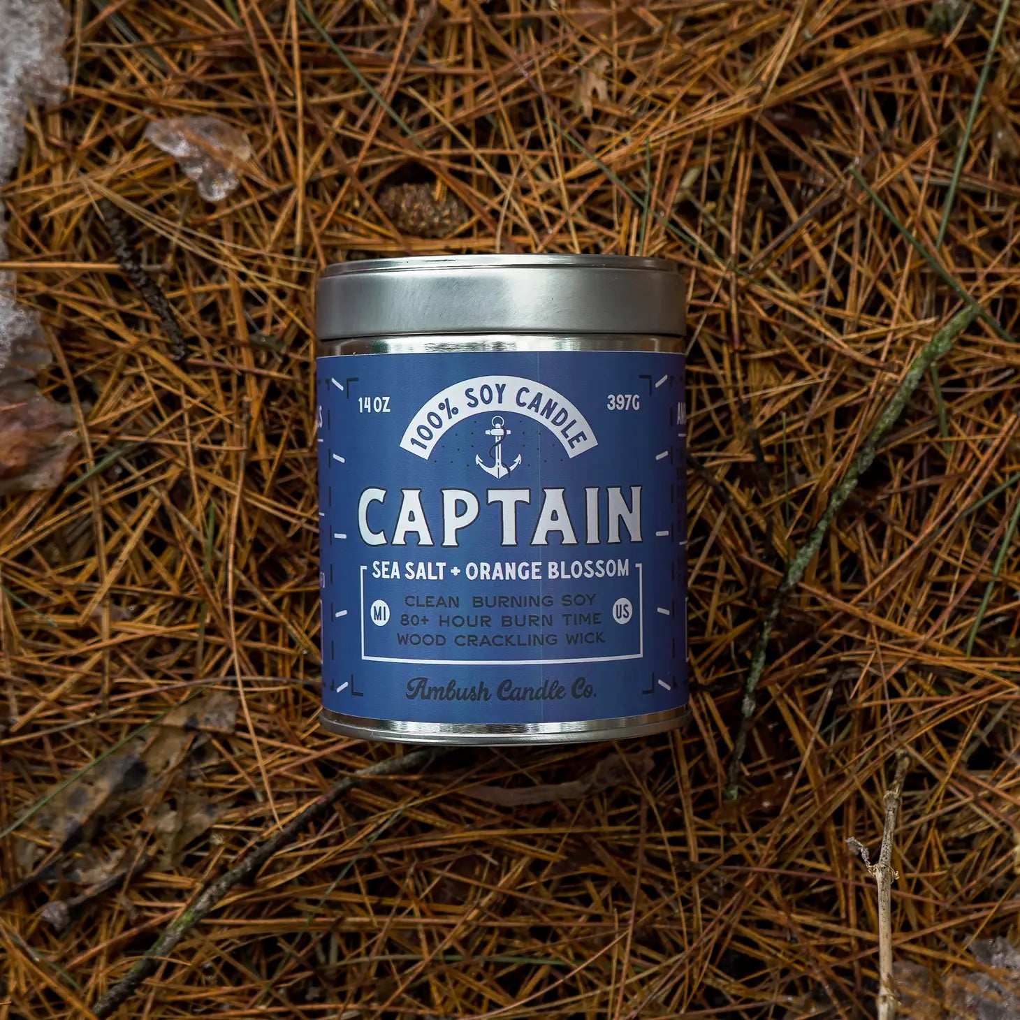 A tin of Ambush Candle Co Captain 14OZ Sea Salt & Orange Blossom Candle, placed on a bed of pine needles.
