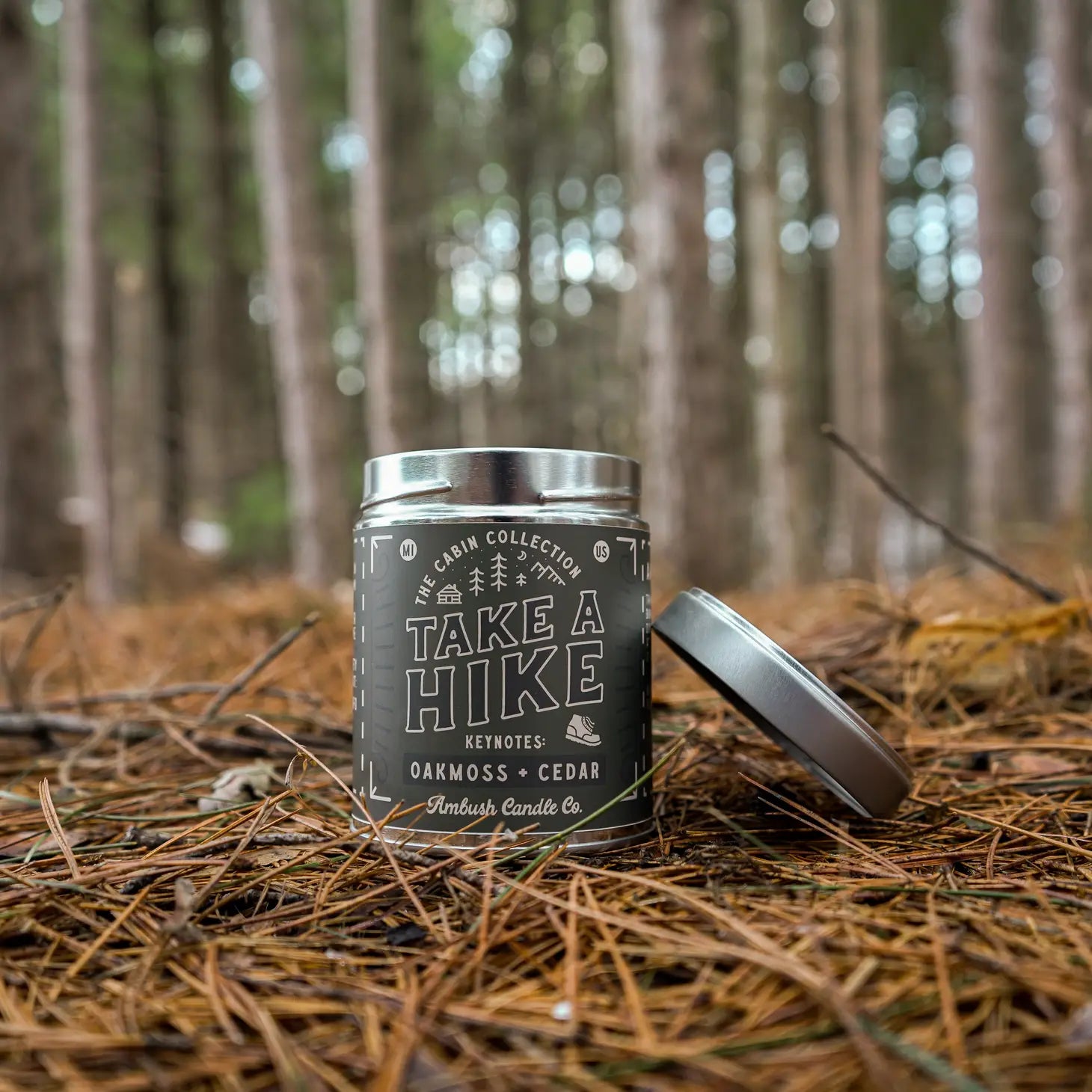 Hand lighting an eco-friendly Ambush Candle Co 14OZ Take A Hike Oakmoss & Cedar Candle in a forest setting, crafted with clean-burning soy wax. It perfectly complements your eco-friendly lifestyle.