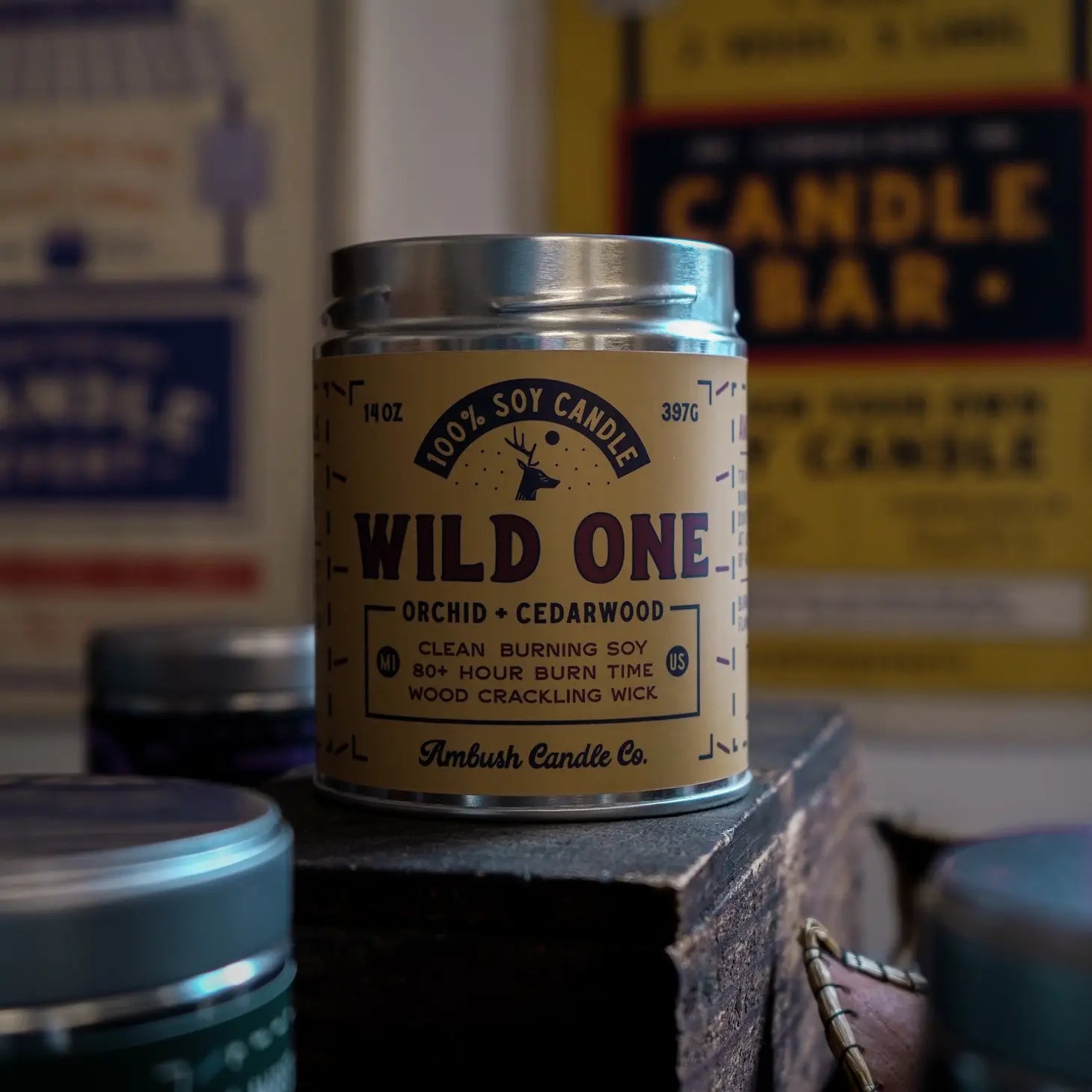 An "Ambush Candle Co 14OZ Wild One Orchid & Cedarwood Candle" crafted from eco-friendly soy wax, housed in a tin and set on a rustic surface. The backdrop of blurred posters enhances the allure of its captivating fragrance.