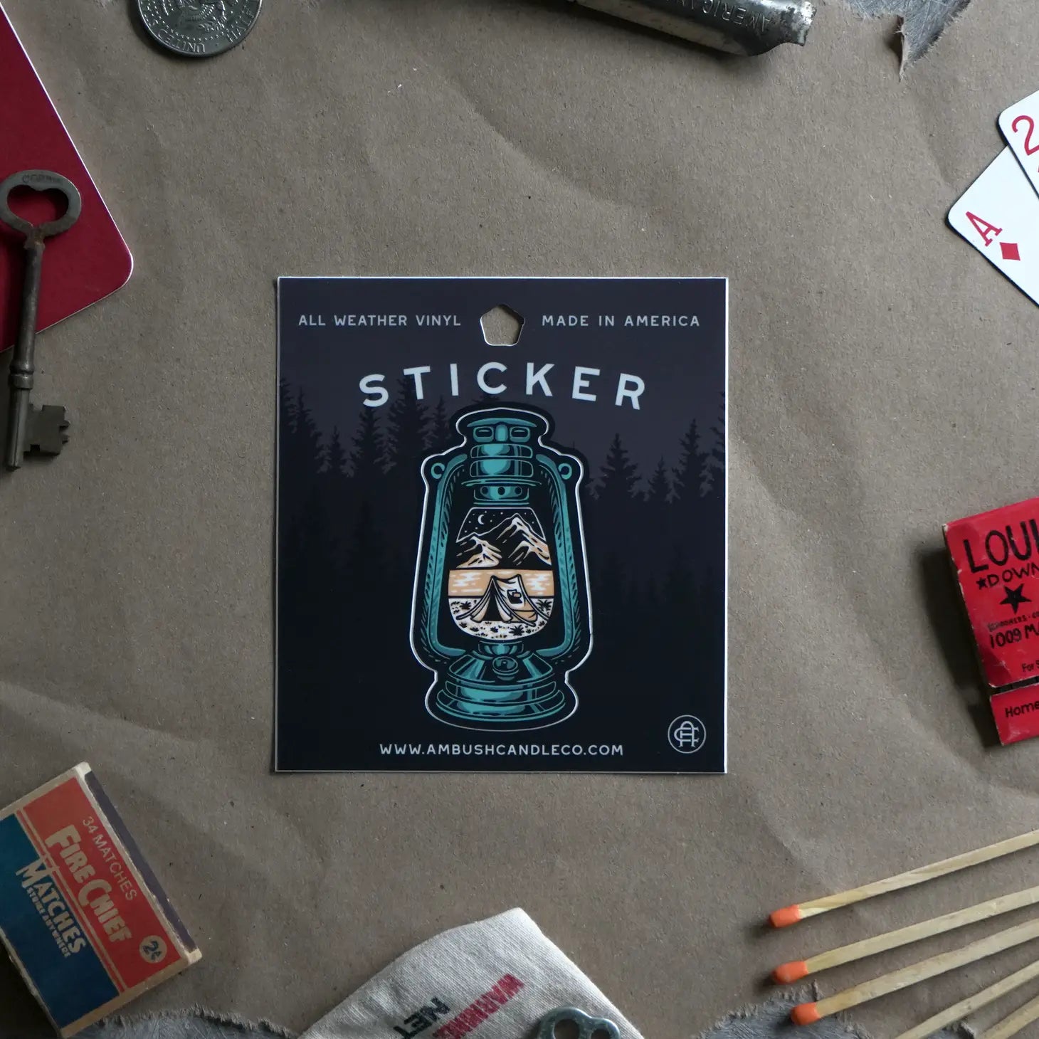 The Ambush Candle Co Lantern Sticker is a weatherproof vinyl sticker featuring a design of a lantern with a tent in a forest scene, encircled by vintage objects on a brown surface. Made in the USA, this removable sticker brings charm to any collection.