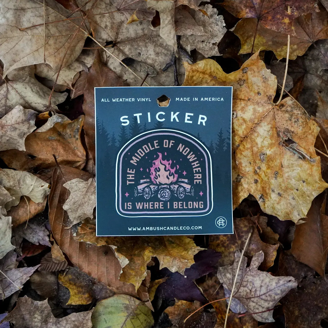 A weatherproof vinyl sticker from Ambush Candle Co, featuring the text "The Middle of No-Where is Where I Belong" and a campfire design, placed on a bed of fallen leaves.
