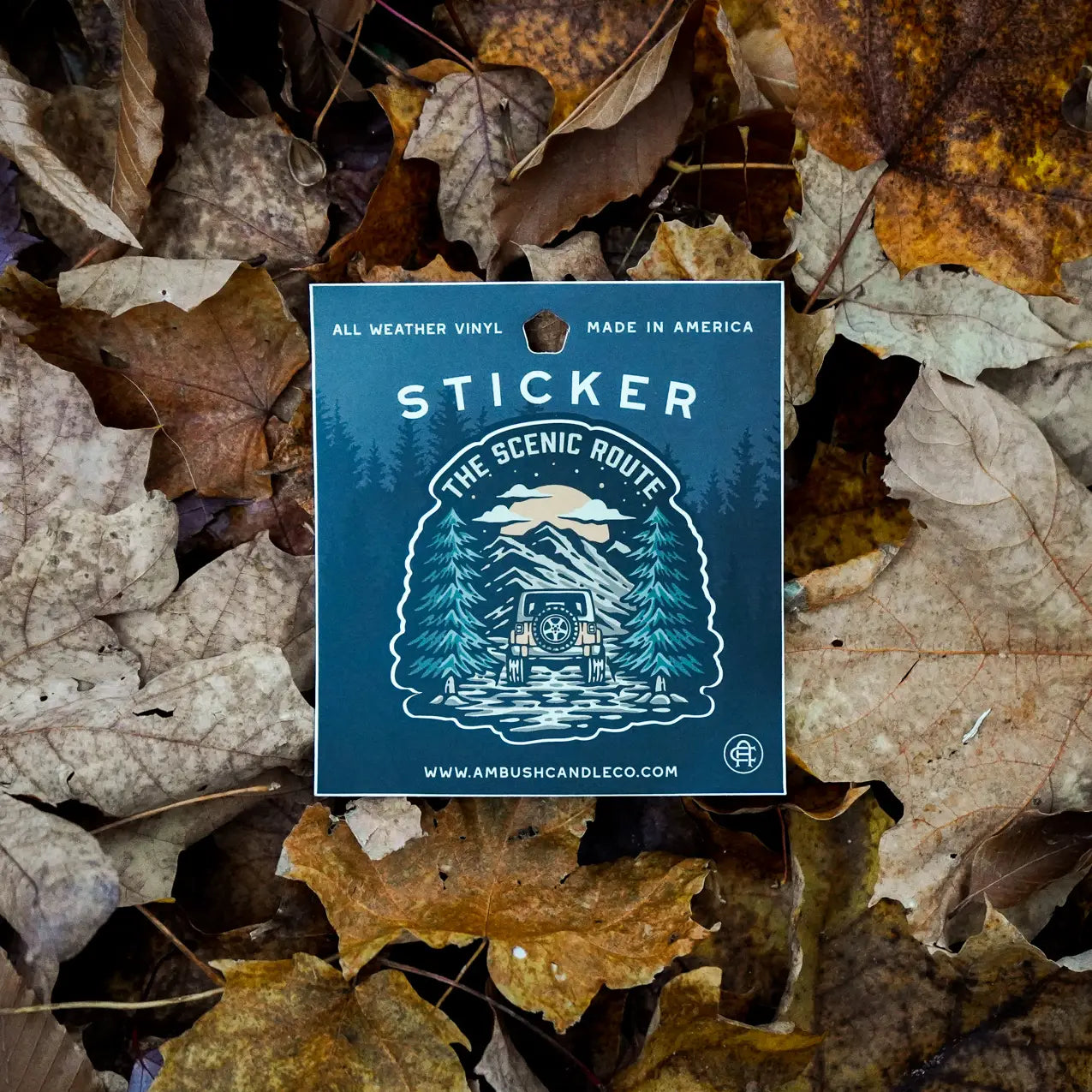 An Ambush Candle Co The Scenic Route Sticker, featuring a weatherproof vinyl design with a snowy cabin scene, is placed on ground covered in dried autumn leaves. Made in the USA, this high-quality sticker is also dishwasher safe.