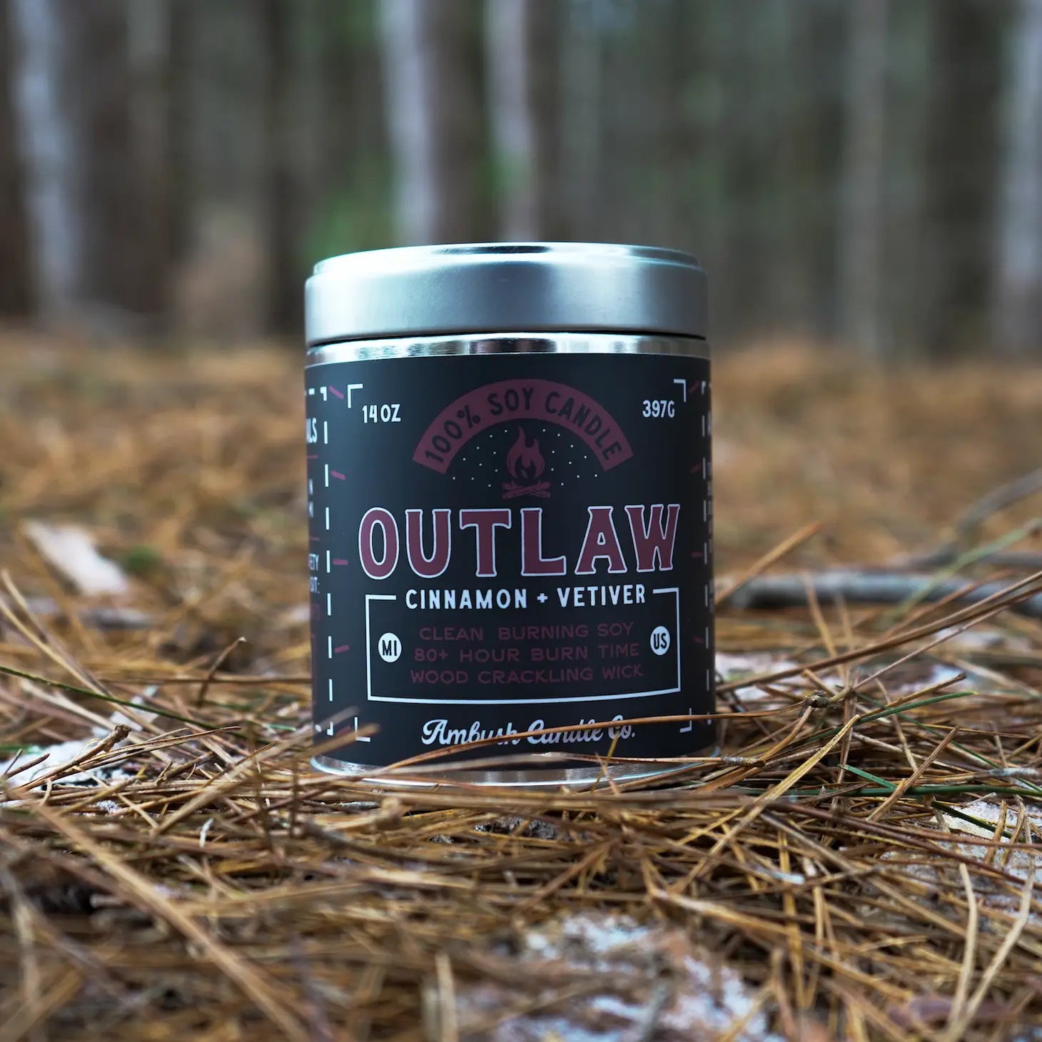A 14oz can of Ambush Candle Co's Cinnamon & Vetiver Outlaw Candle, known for its eco-friendly nature, stands on pine needles in a forest setting and offers an impressive 80 hours of burn time.