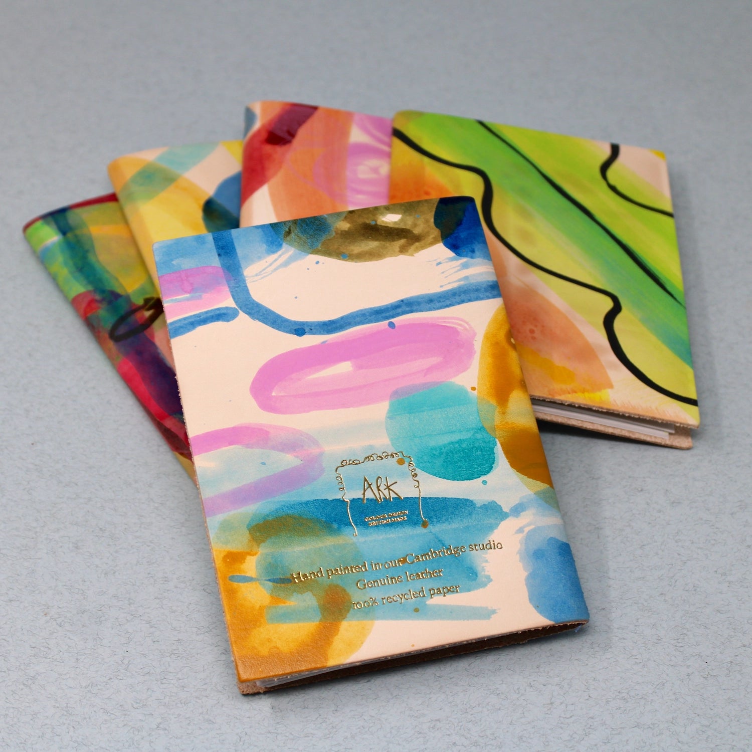 Handpainted A6 leather sketchbooks by Four Ark Colour Design stacked neatly on a light gray surface, premium art supplies.