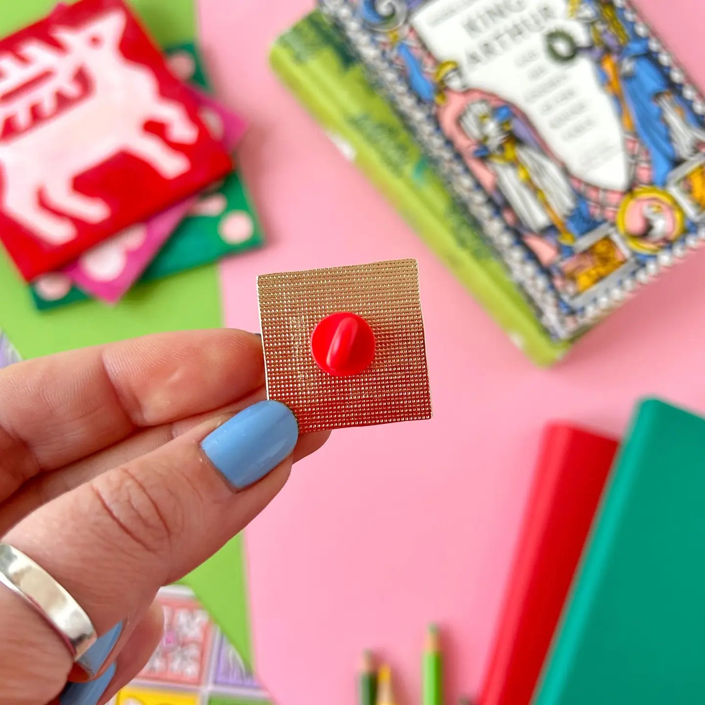 Blue-nailed hand holding Stacey McEvoy Caunt Lion Medieval Tile Pin with books, cards on pink background; enamel pin art.