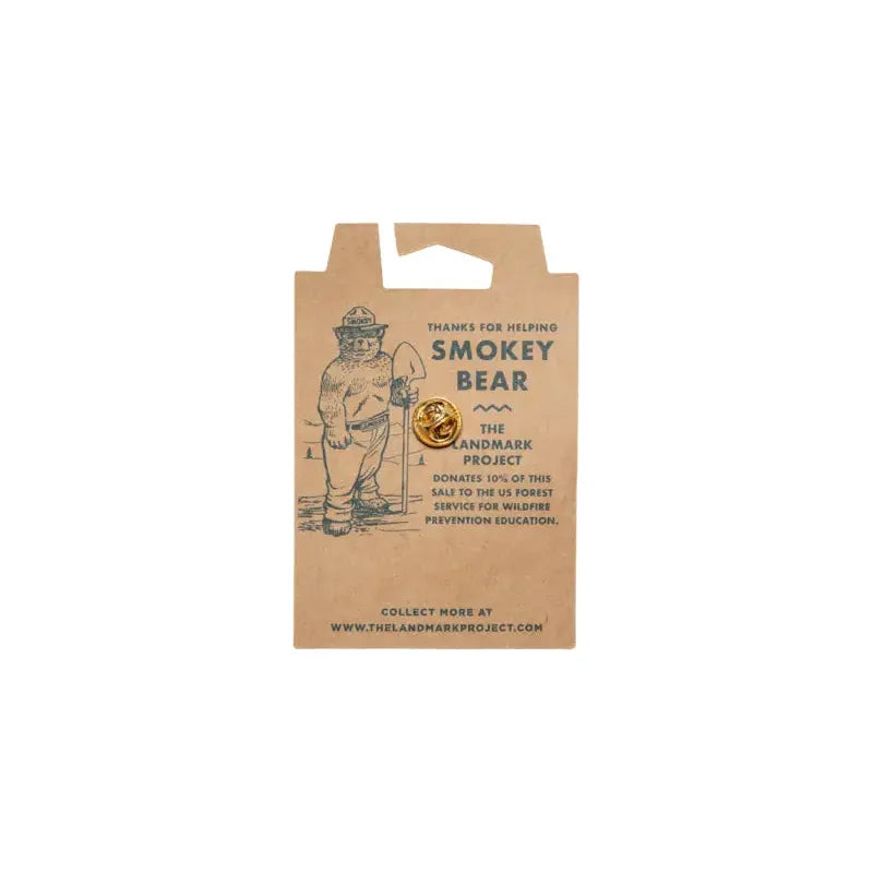 The Landmark Project Smokey Enamel Pin - Smokey Logo is packaged on a card showcasing a drawing of Smokey Bear along with text promoting wildfire prevention and The Landmark Project, all encased in sustainable materials.