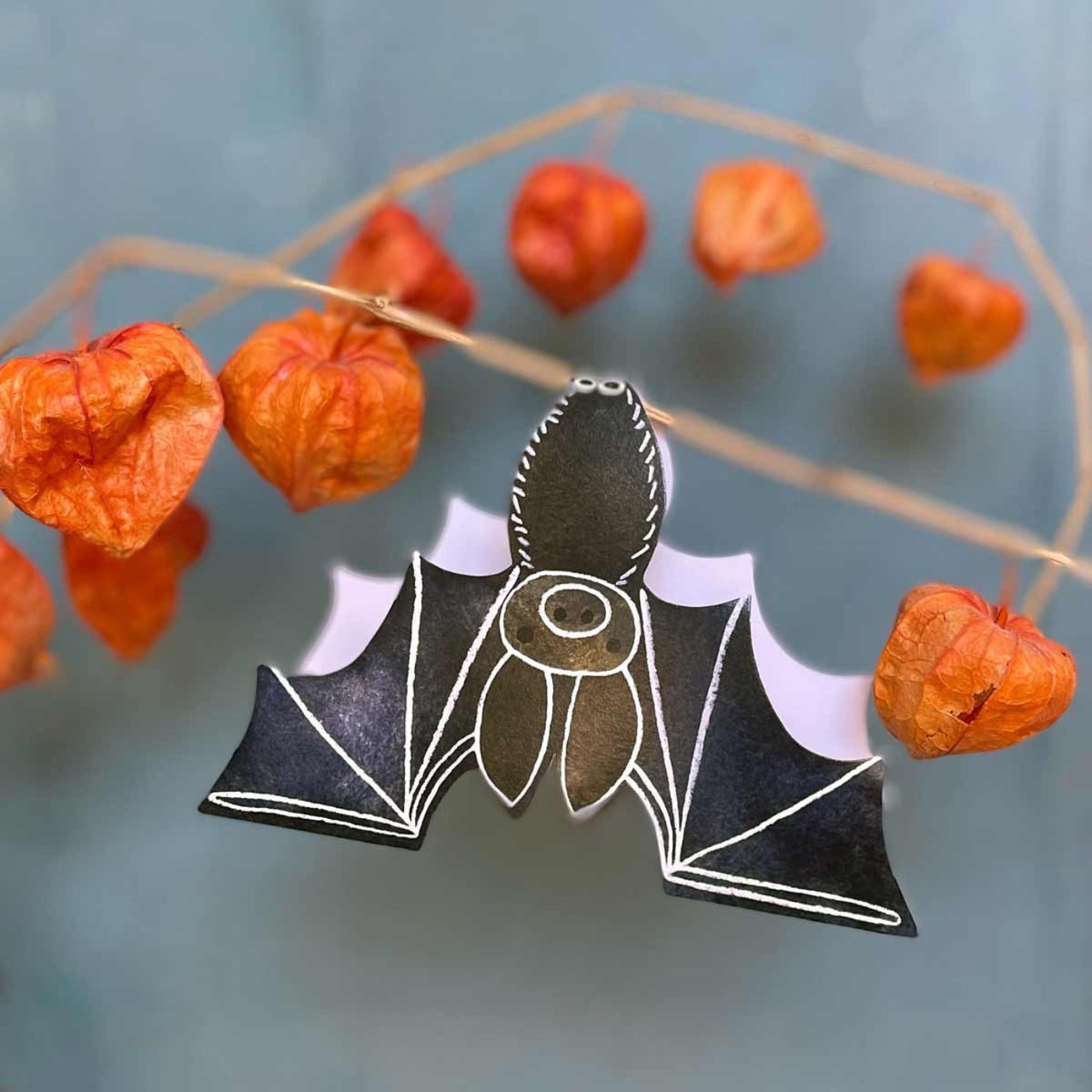 A card in the shape of a illustrated bat with dried Japanese Lantern plant around it