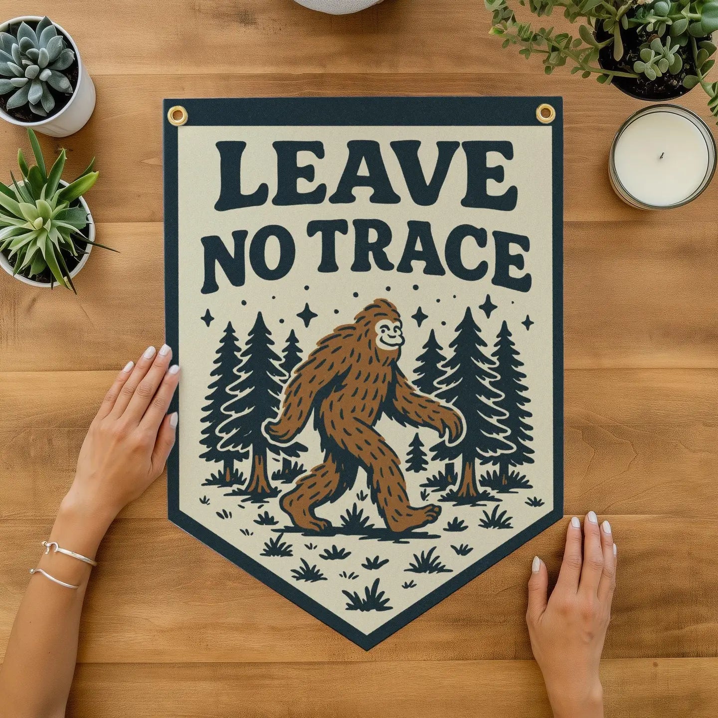 Person displays Benoit’s Design Co Leave No Trace Bigfoot Banner Pennant—perfect for rustic cabin wall décor with pine trees.