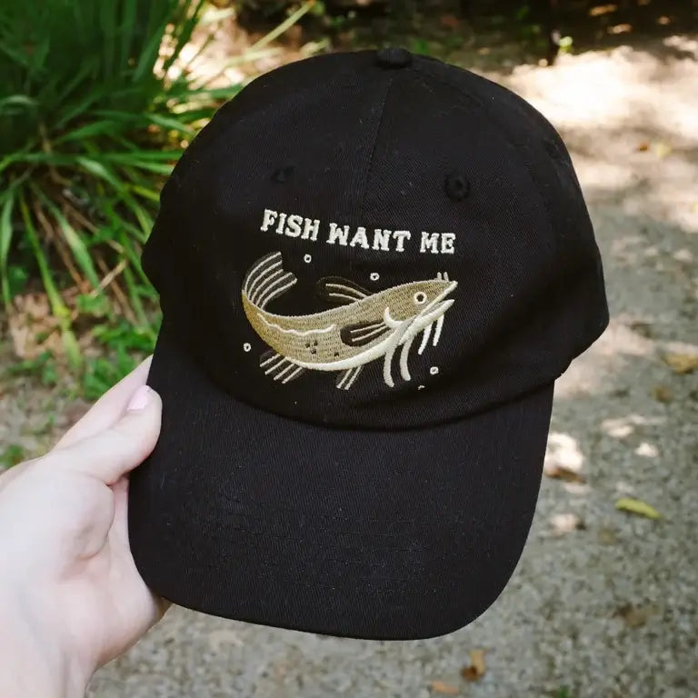 Black cap with green and beige cat fish graphic and 'Fish Want Me' text held outdoors.