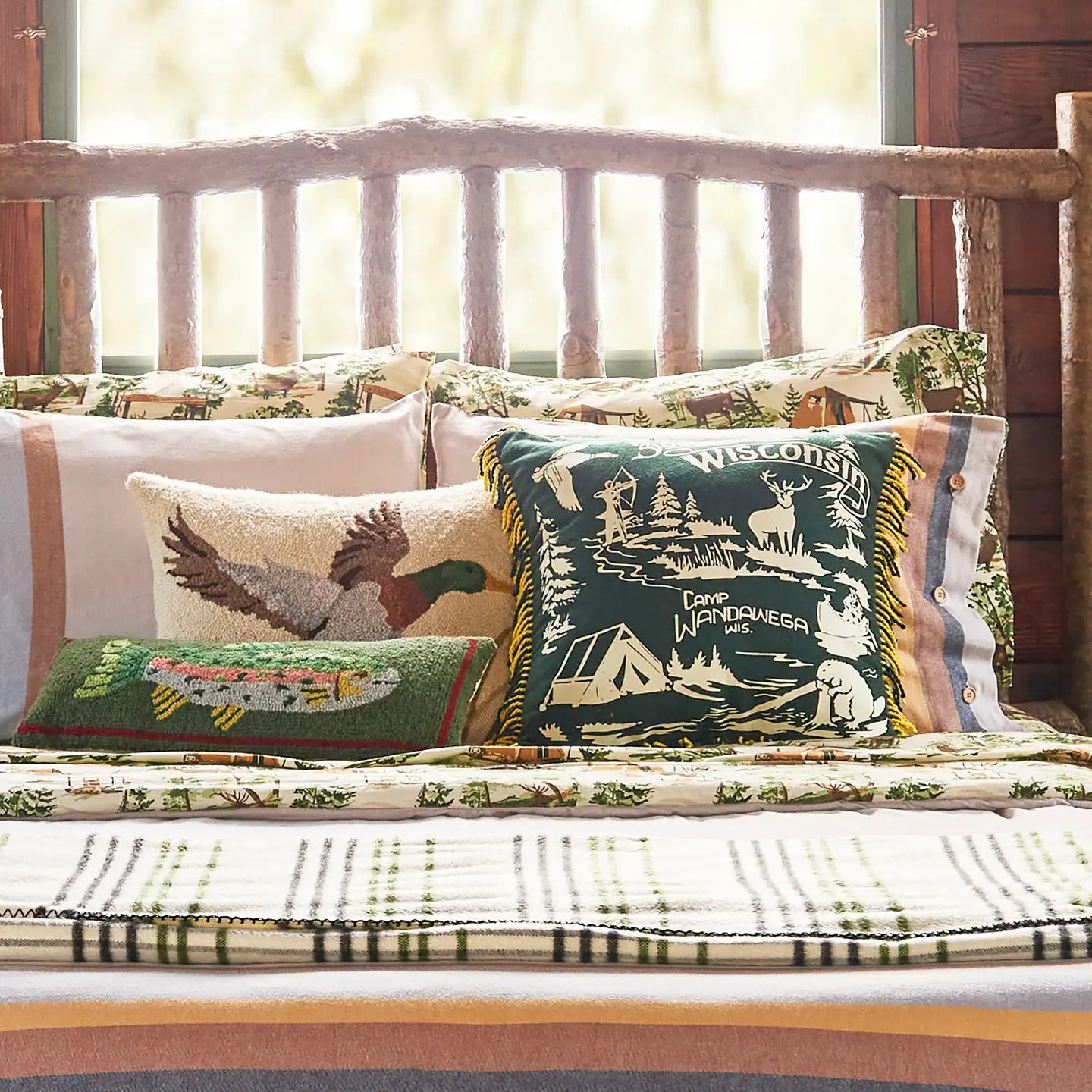 Rustic wooden bed with plaid bedding and Camp Wandawega Trout Hook Pillow for cozy lodge-style bedroom decor.