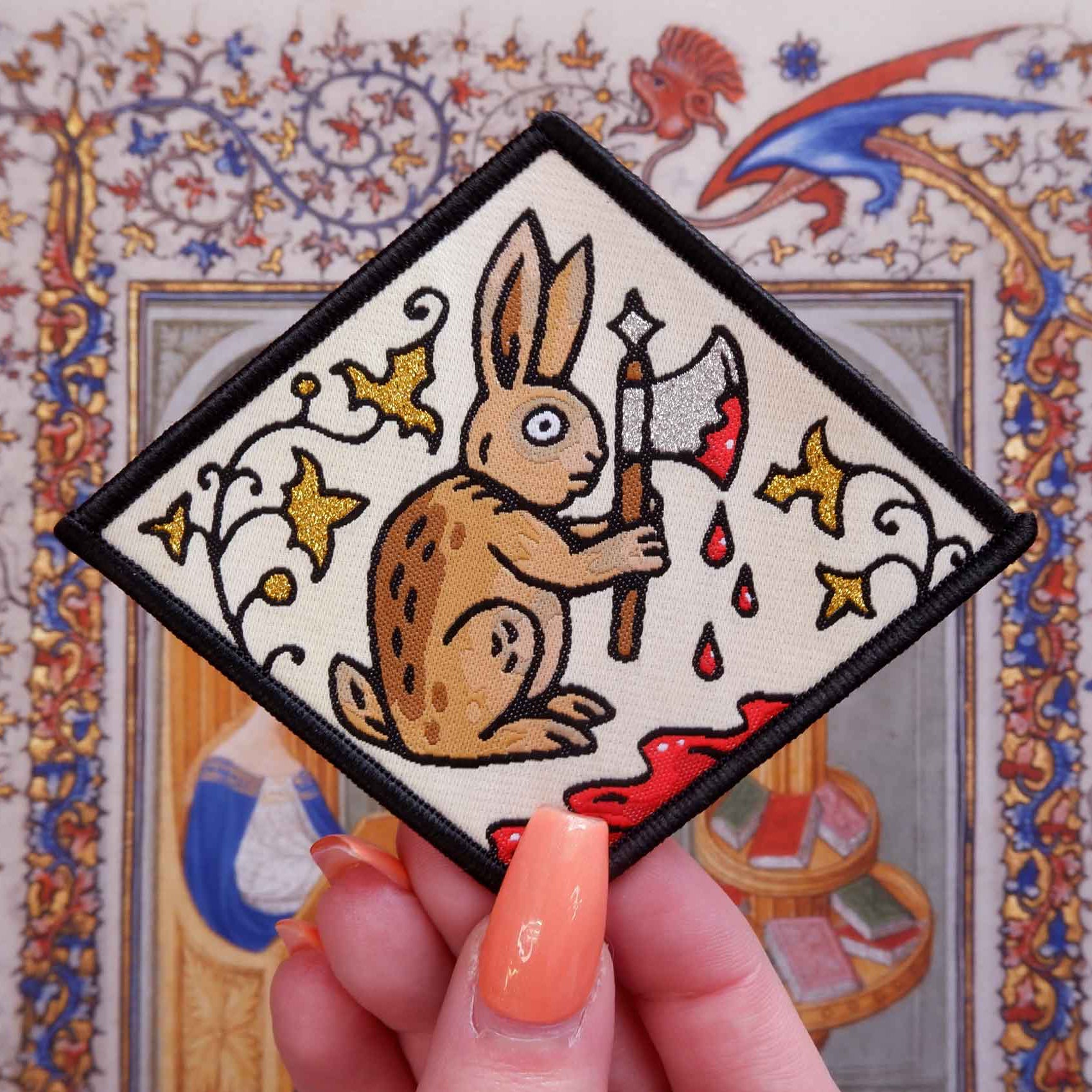 Hand holding Cat Coven Medieval Rabbit Patch: rabbit with bloody axe, medieval art style, unique woven accessory for jackets.