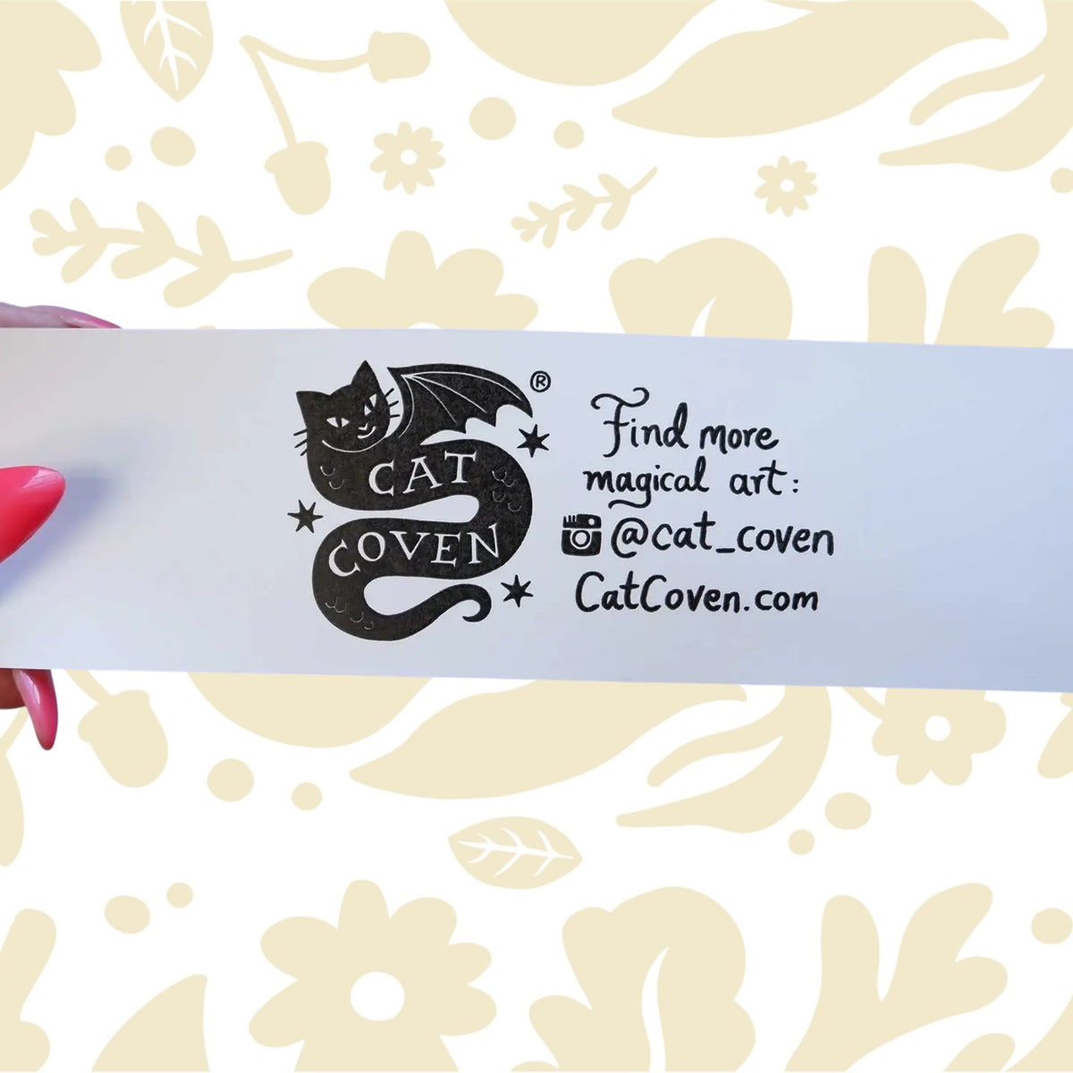 Hand holding Cat Coven Sphinx vs Dragon holographic sticker card with logo, website, IG on floral background.
