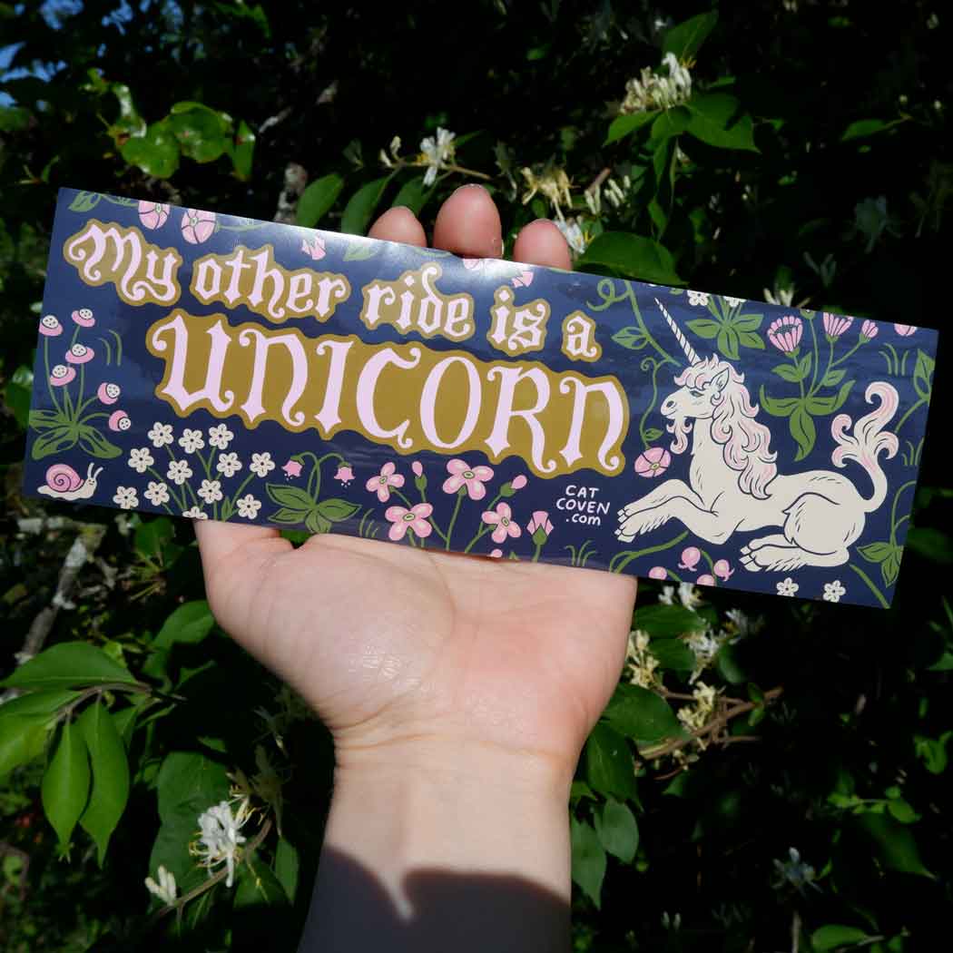 Hand displaying Cat Coven "My Other Ride Is A Unicorn" bumper sticker with magical, floral art for fantasy car accessory lovers.