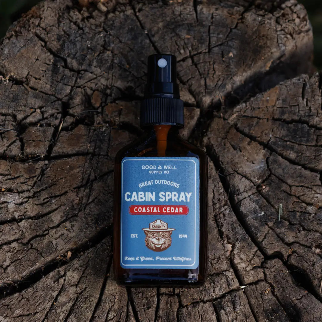 Good & Well Supply Co Coastal Cedar Smokey Bear Cabin Spray bottle on tree stump outdoors, rustic natural home scent decor.
