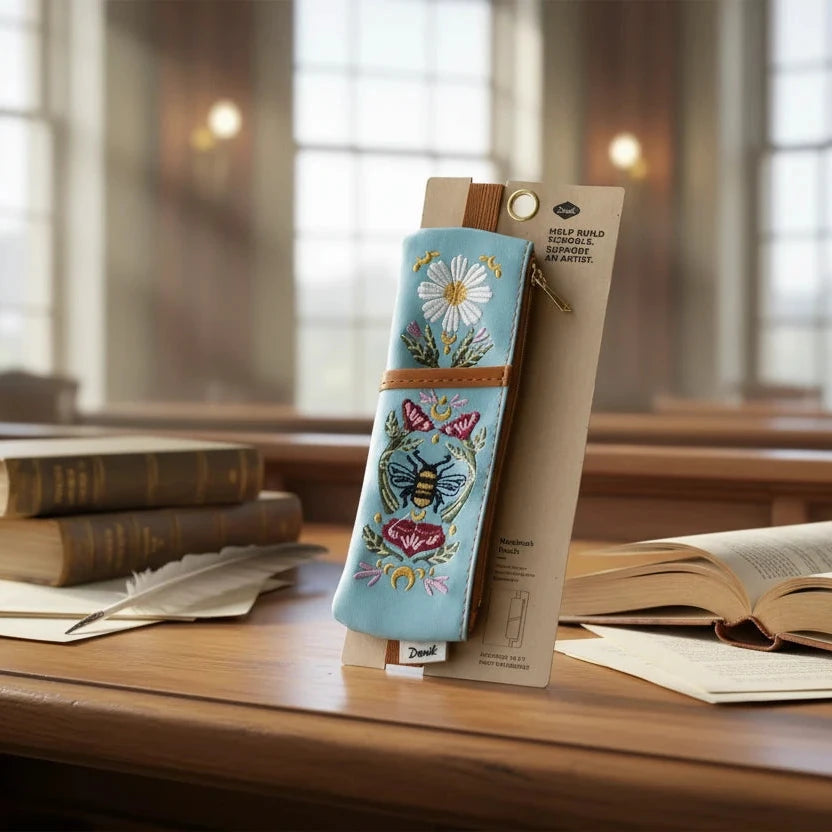 Blue Denik embroidered bee vegan leather pouch with floral print on brown notebook by window; stylish accessory organizer.