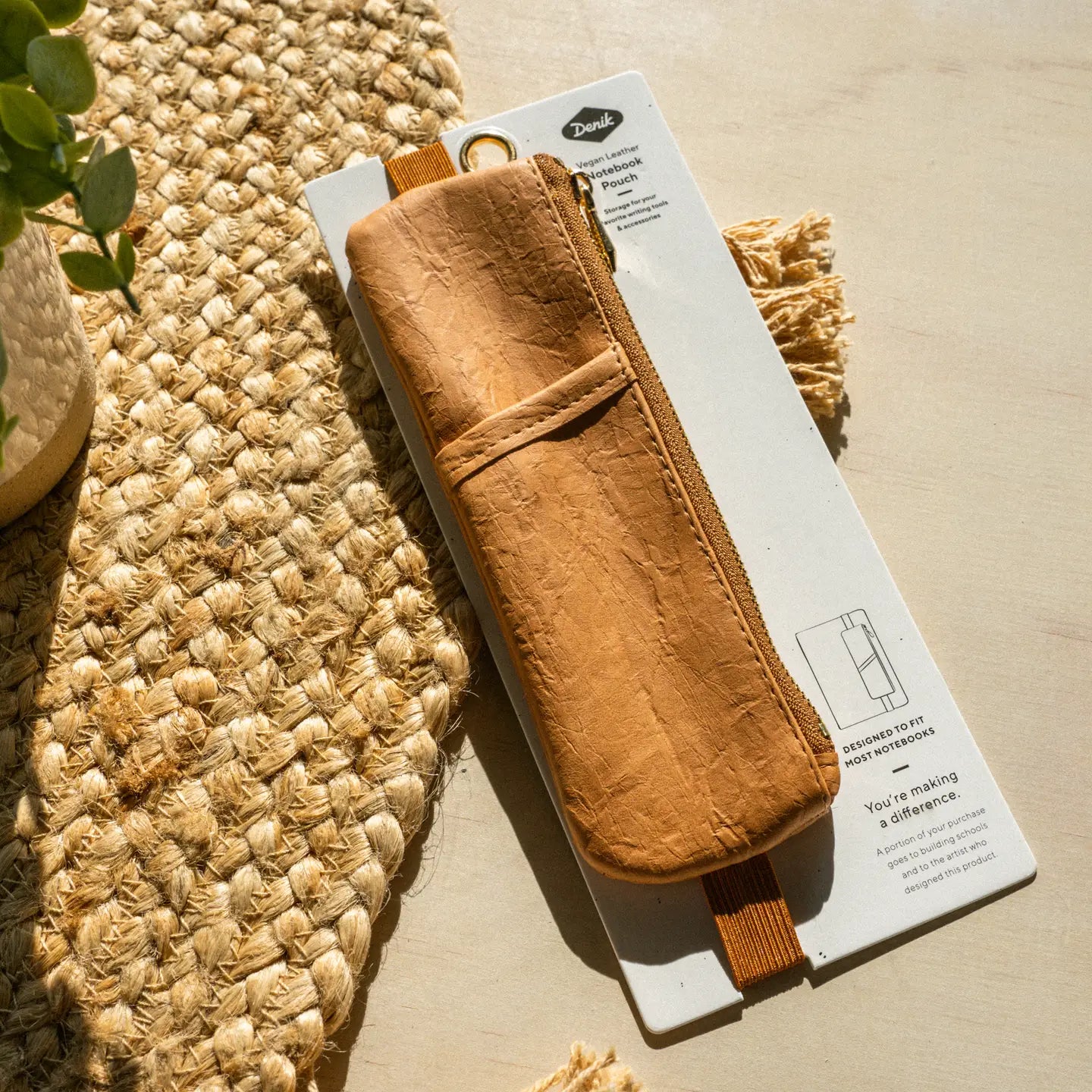 Denik Caramel Crinkle Vegan Leather Notebook Pouch with zipper on cardboard, next to a woven mat and houseplant for desk decor.