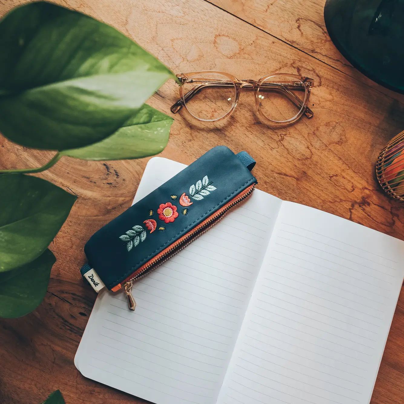 Denik Envelyn blue embroidered folk flower vegan leather pouch sits on a desk, paired with a notebook and eyeglasses for work.