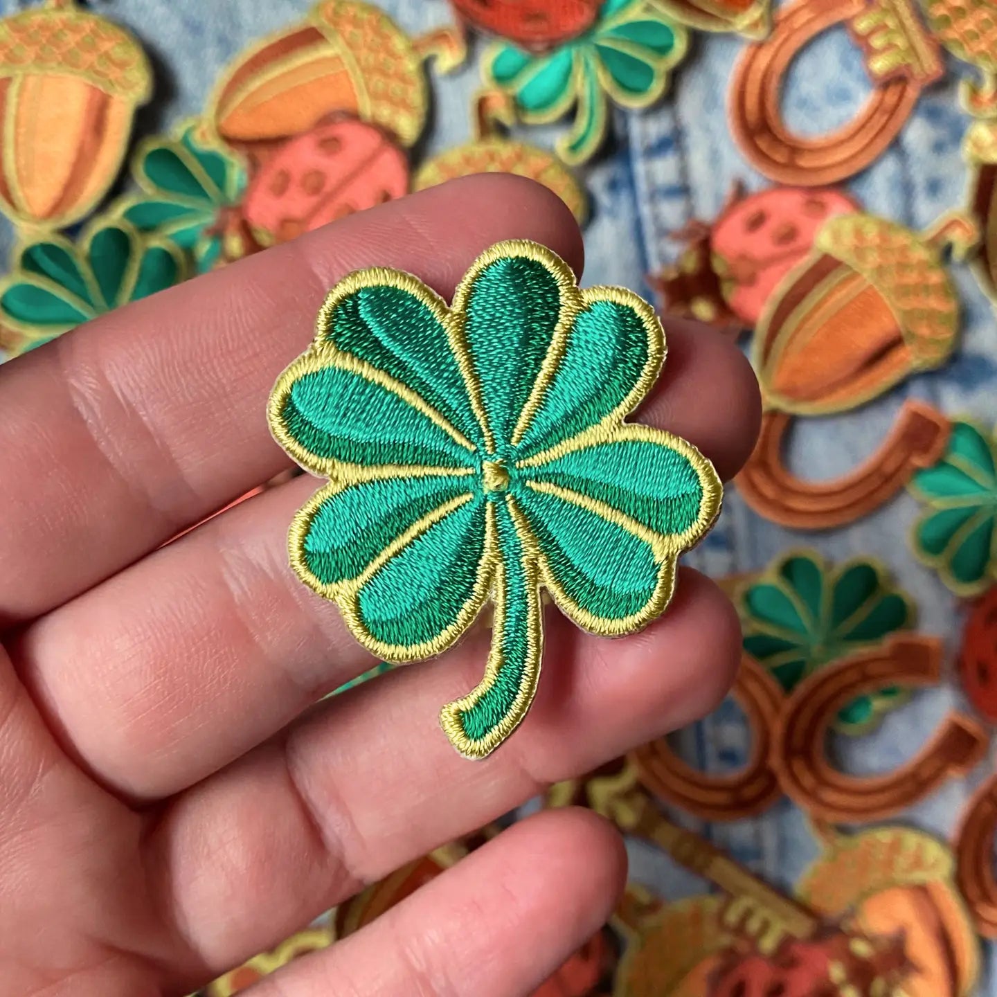 Hand holding Kate O’Hara Good Luck Embroidered 5 Patch Set; lucky acorns and horseshoes showcased in background for luck.