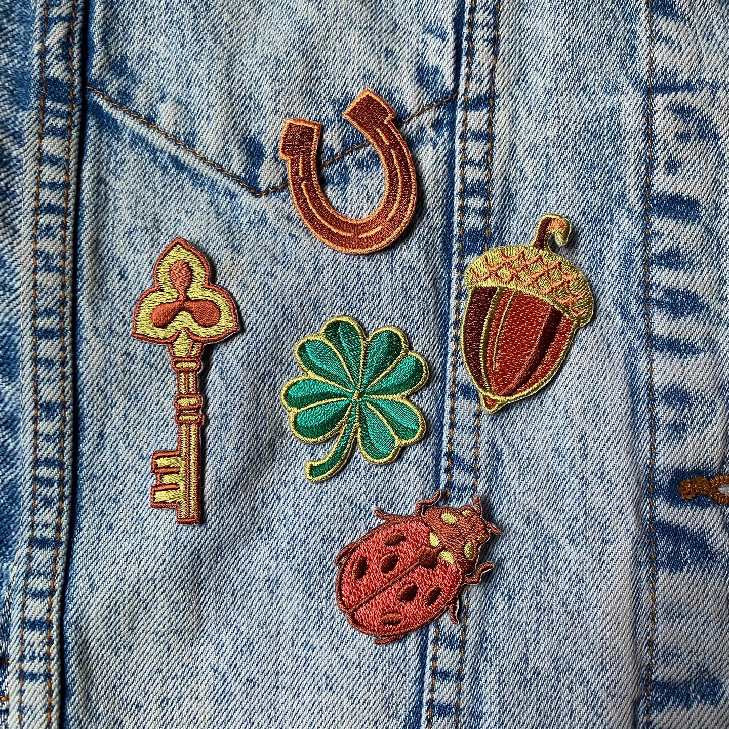 Kate O’Hara Good Luck embroidered patch set—key, horseshoe, acorn, clover & ladybug on blue denim jacket pocket.