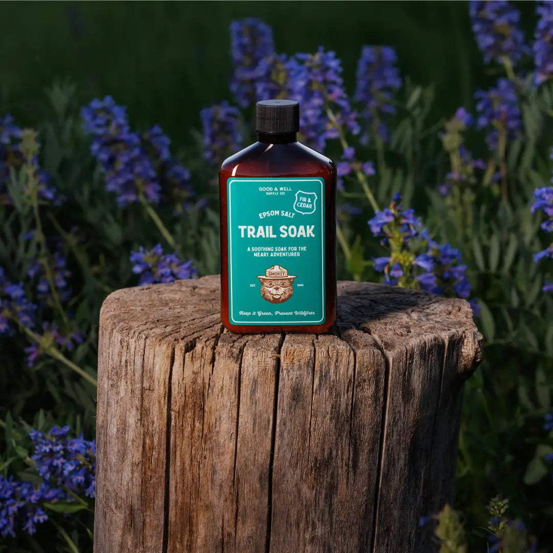Good & Well Supply Co Smokey Bear Trail Soak Fir & Cedar bottle on tree stump, surrounded by lush grass and wildflowers outdoors.