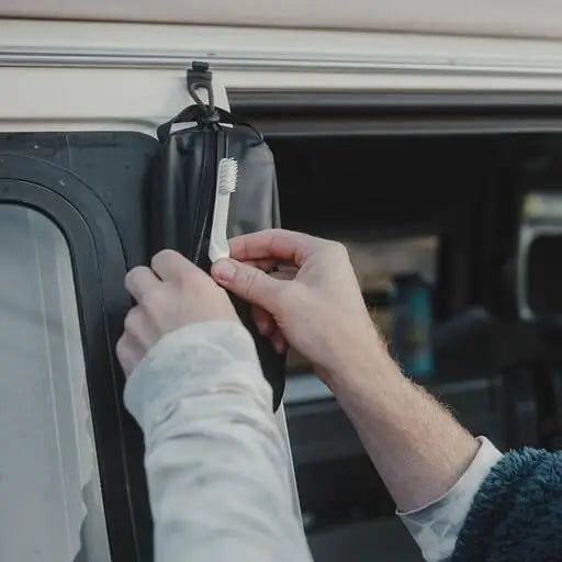Traveler packing toothbrush into Gravel Outdoor Explorer Mini™ Toiletry Bag hanging at car window—perfect for outdoor trips.