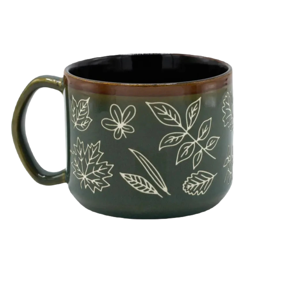 A Cognitive Surplus Pressed Leaves Hand Carved 15oz Ceramic Mug in dark green, adorned with white floral and leaf designs in a hand-carved botanical pattern and a brown rim.