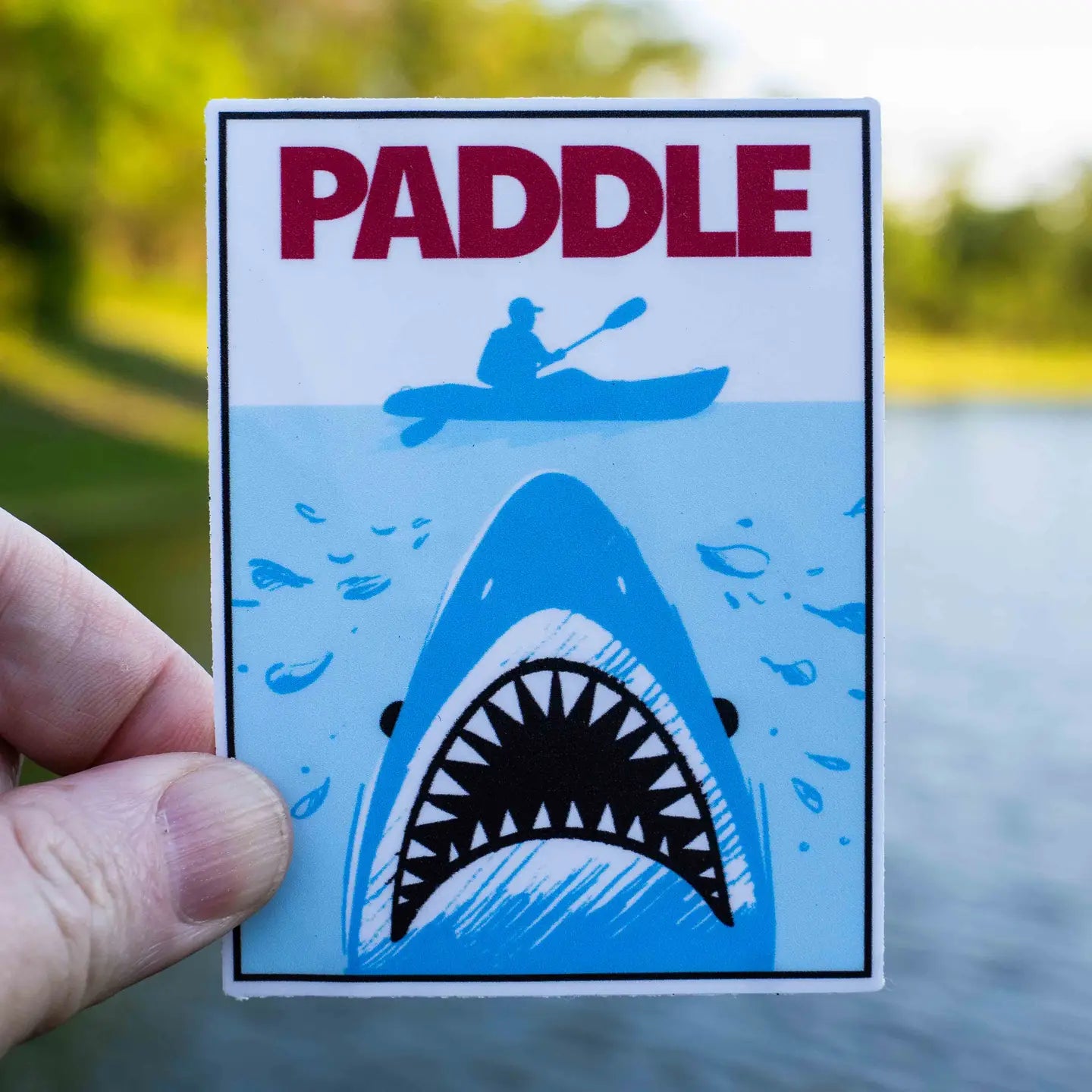 Hand holds Hey Bear Outdoors Jaws Kayaking Paddle Sticker, kayaker over shark, bold red PADDLE—ideal kayaking gift.