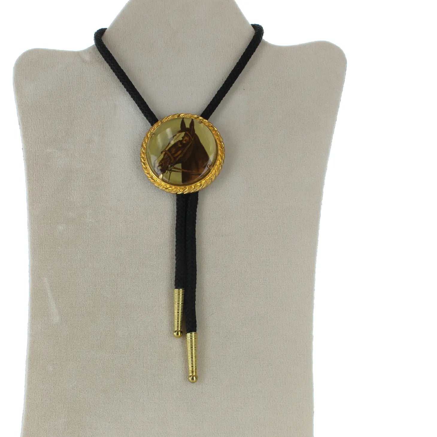 The Forager Provisions Horse Portrait Bolo Tie is hand-made and displayed on a white stand, featuring a large, gold and brown decorative slide. It also includes black braided cords with gold tips and an adjustable cord for a perfect fit.