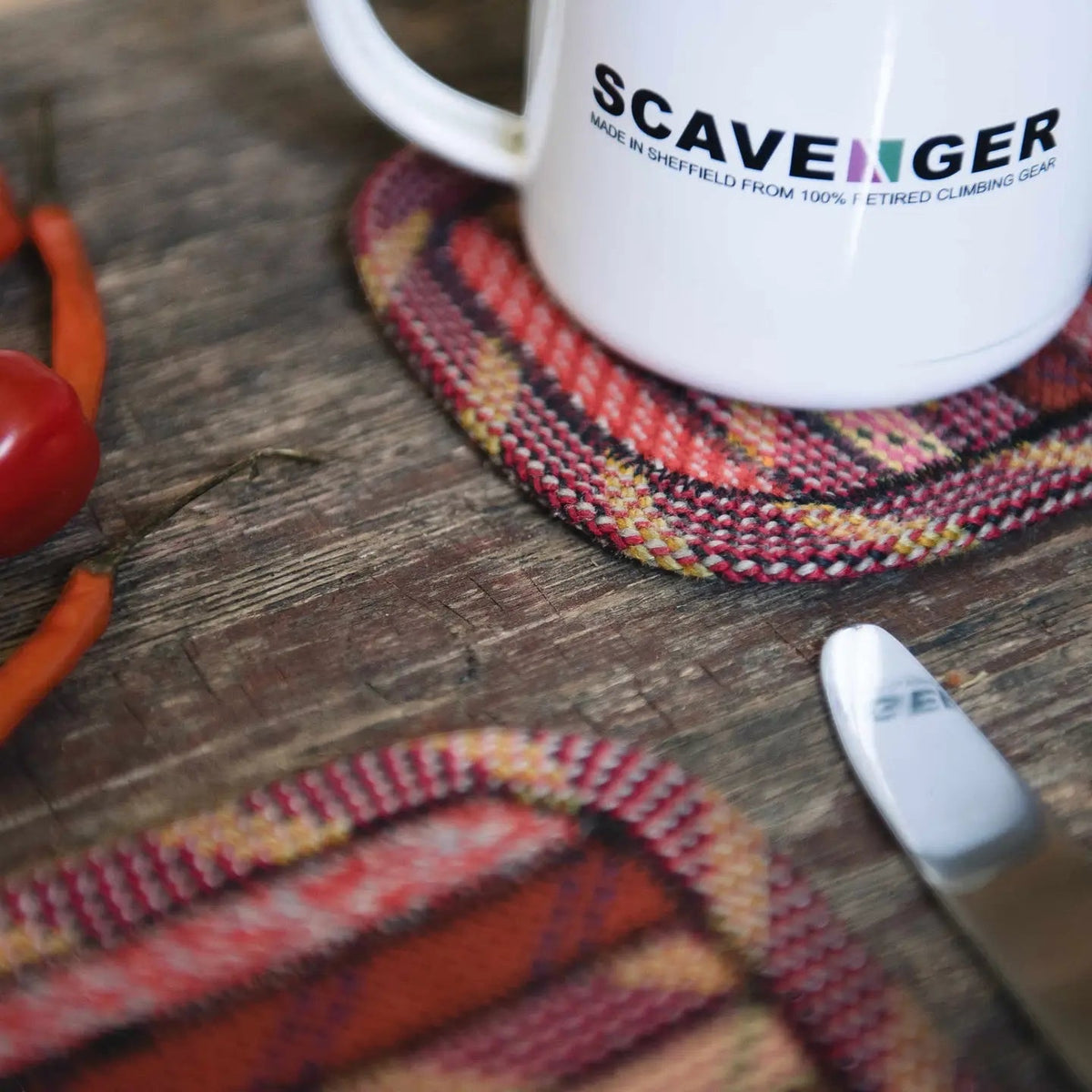 White mug on Scavenger Upcycled Fire Climbing Rope Coaster with knife, chili peppers, tomato on rustic wood background.