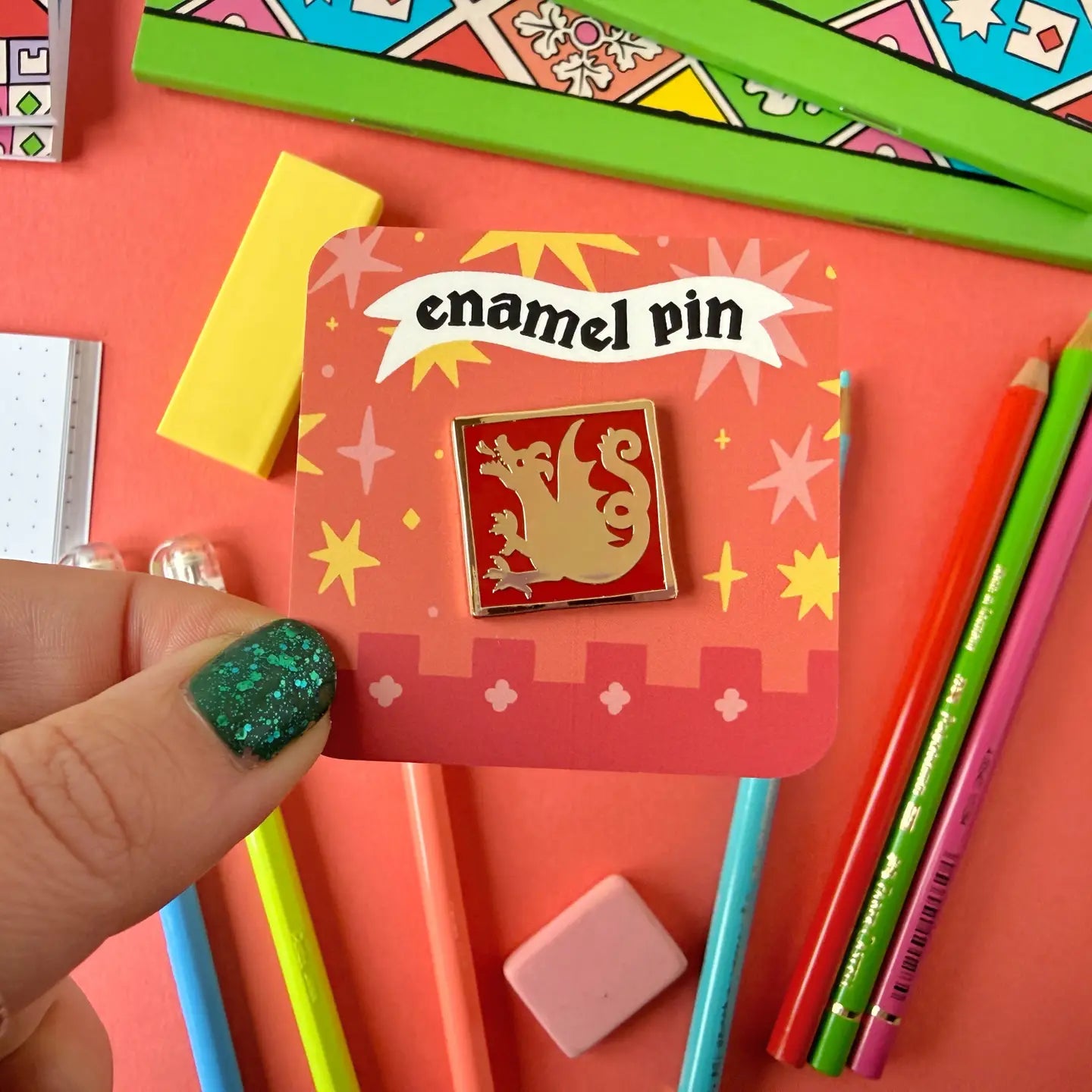 Hand displays Stacey McEvoy Caunt Medieval Mini Dragon Enamel Pin on card, surrounded by unique stationery for collectors.