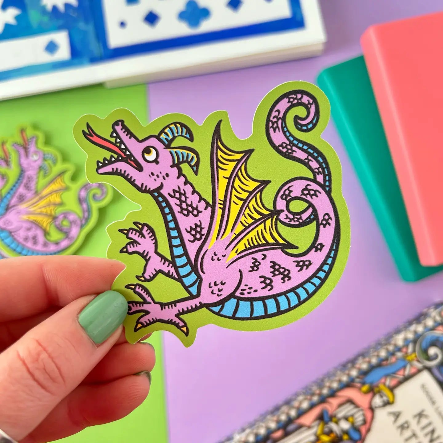 Hand displaying Stacey McEvoy Caunt Dragon Medieval Marginalia Vinyl Sticker with stationery, notebooks, and cards in background.