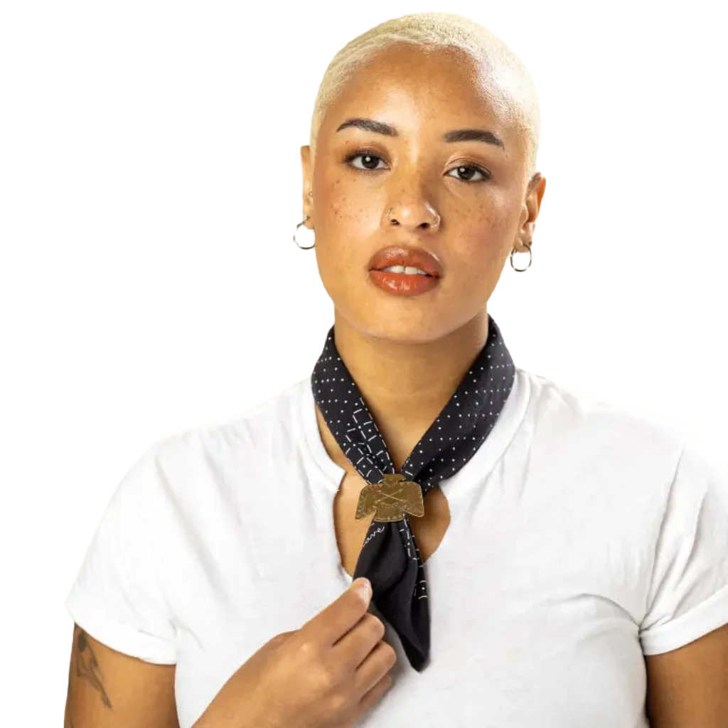 A green scarf with white geometric patterns, styled in a loop and secured by the Jenni Earle Brass Thunderbird Handmade Bandana Slide, brings a touch of western style.