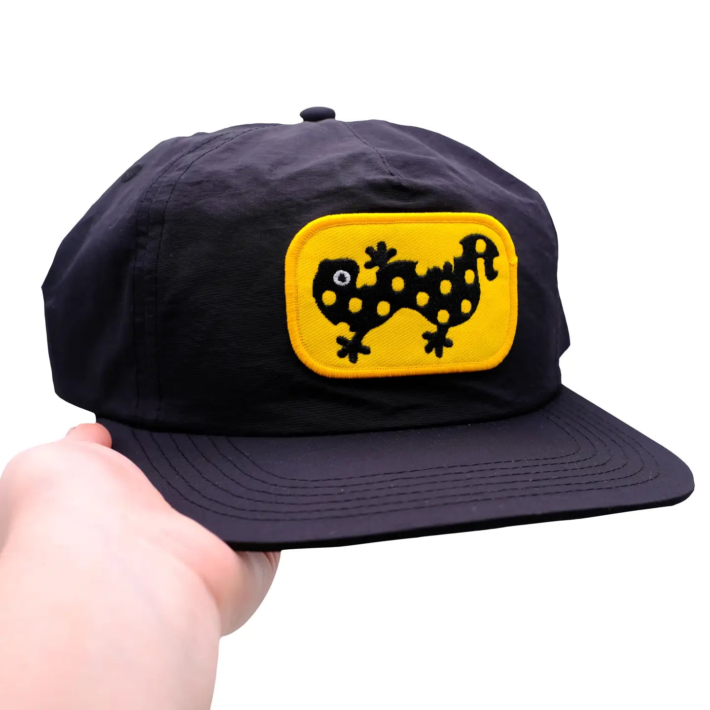 Hand holding Camp Mustelid cap with eye-catching yellow patch, black spotted salamander lizard embroidered on the front.
