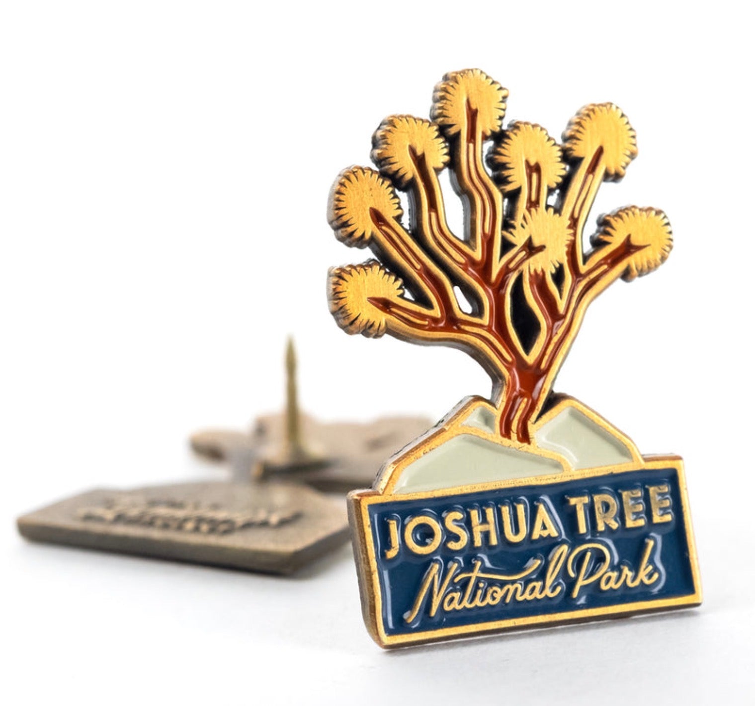 The Landmark Project Joshua Tree enamel pin shows a tree, mountains, and a blurred pin—perfect souvenir for nature lovers.