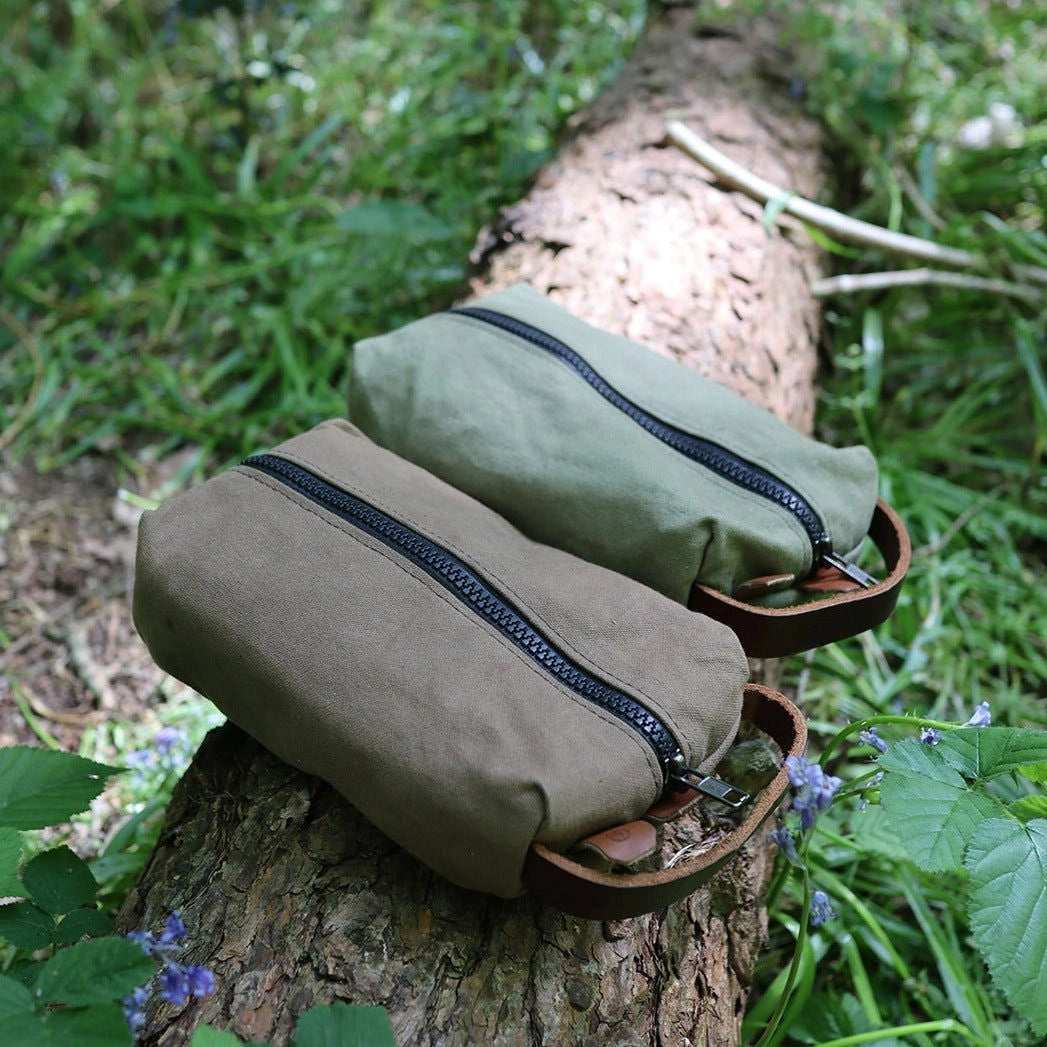 Forager Provisions LTD green and brown canvas toiletry bags with YKK zippers resting on a log in a forest setting.