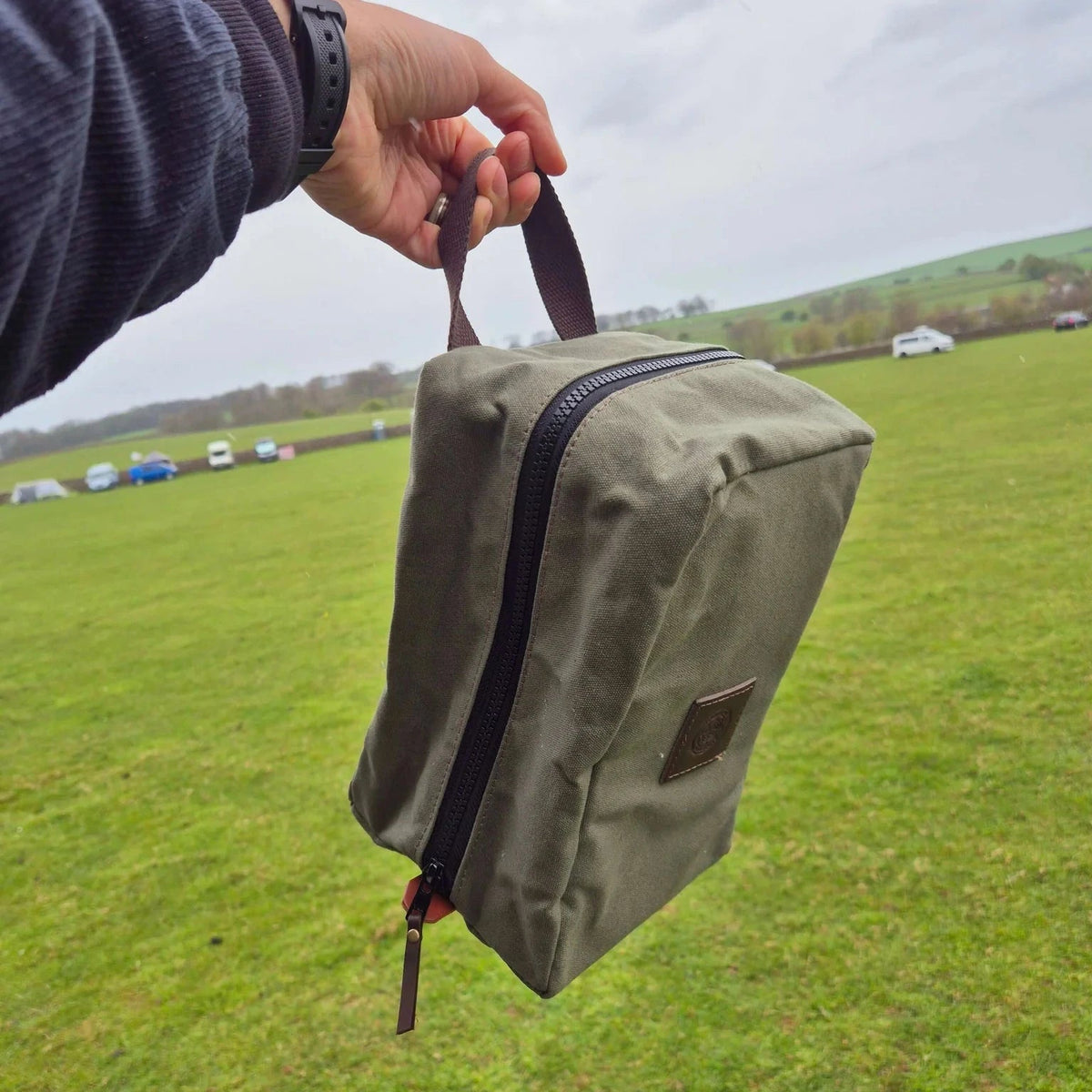 Forager Provisions LTD green waxed canvas map and storage case held by a handle against a grassy outdoor landscape.