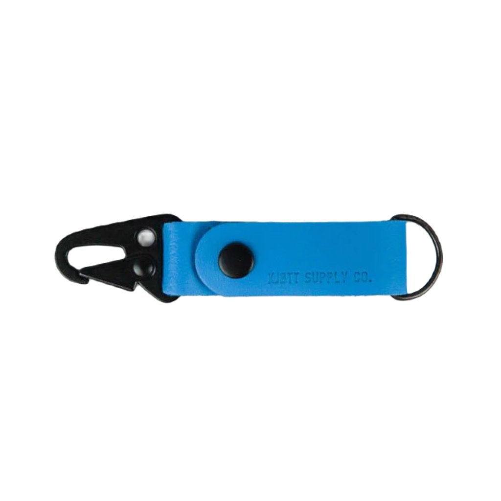 A blue Kjøtt Supply Co Key Fob, featuring a black metal hook, a black button, and the label "KOTT SUPPLY CO." against a white background, crafted from premium leather and backed by a lifetime warranty.