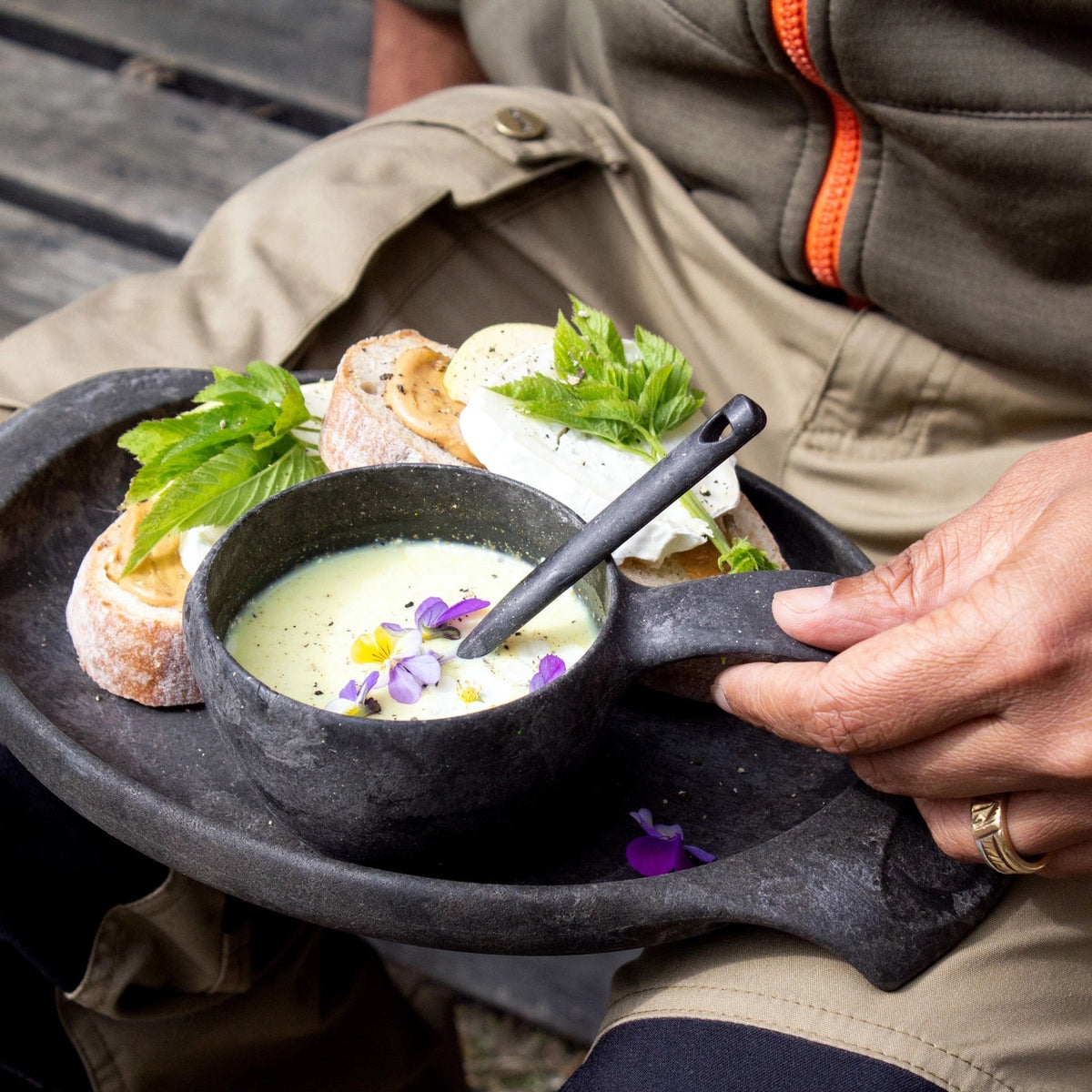 Hand holding Kupilka 44 Large Plate in Brown with gourmet soup, edible flowers, bread, cheese, and fresh herbs—outdoor food serving.