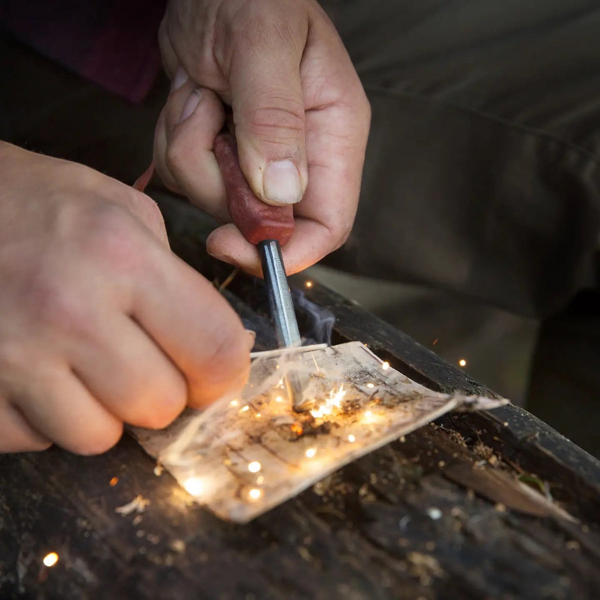 Hands ignite tinder on wood using the Kupilka Firesteel 8 Brown and striker—outdoor survival fire starter in action.